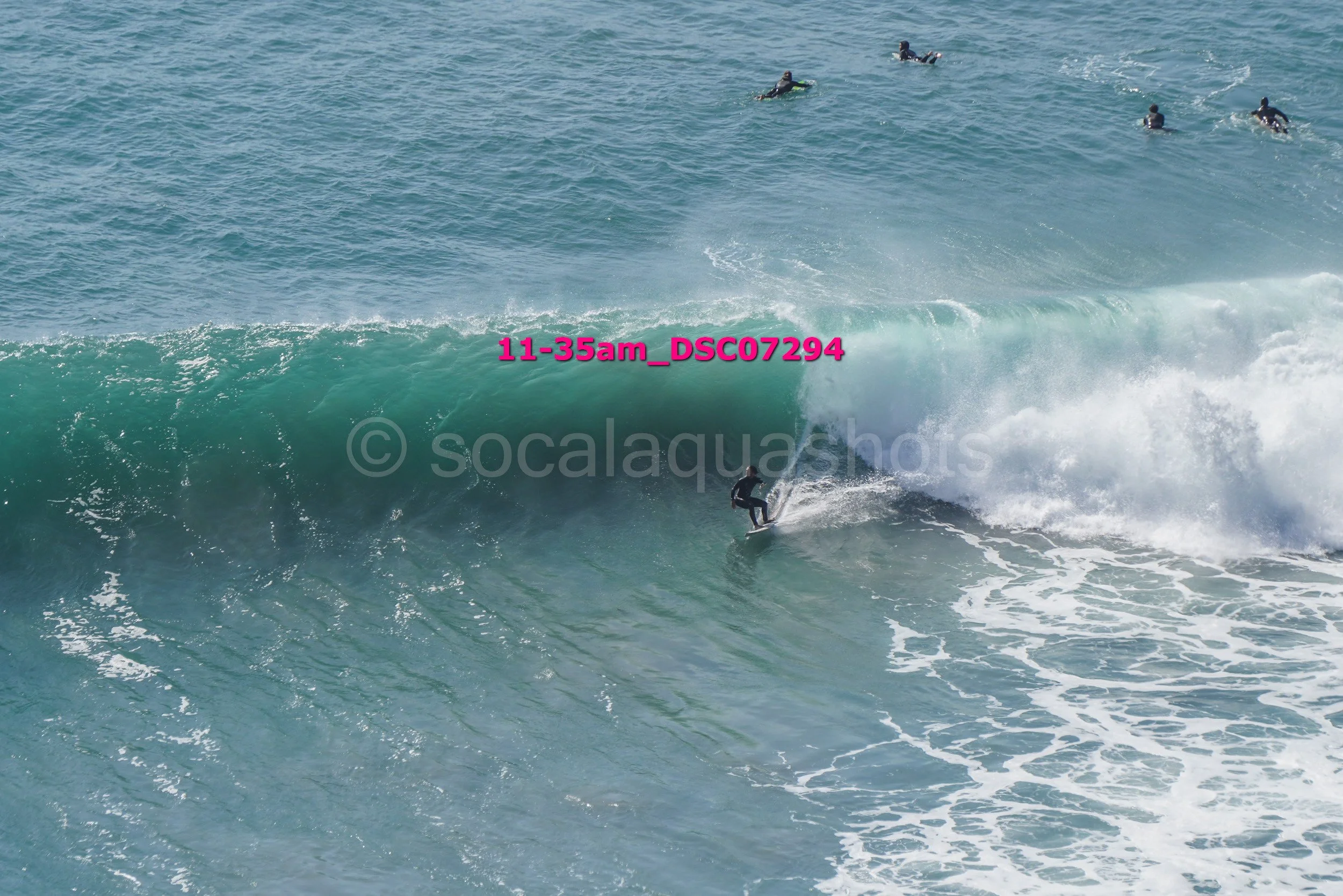 Surfer riding a large wave in the ocean with several other surfers in the water nearby.
