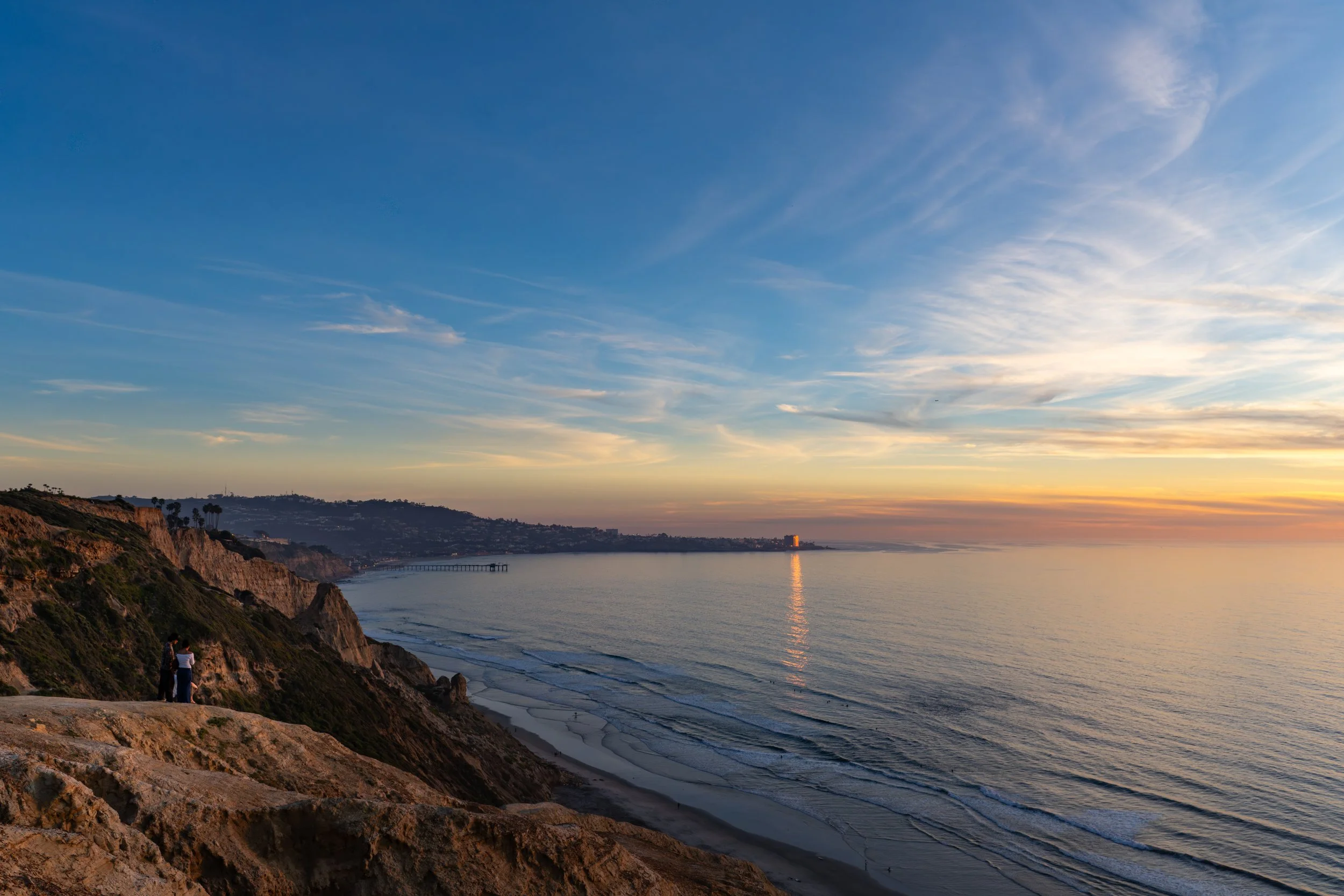 Sunset over a coastal cliffside with two people standing on the rocks, overlooking the ocean with calm waves and a distant pier.