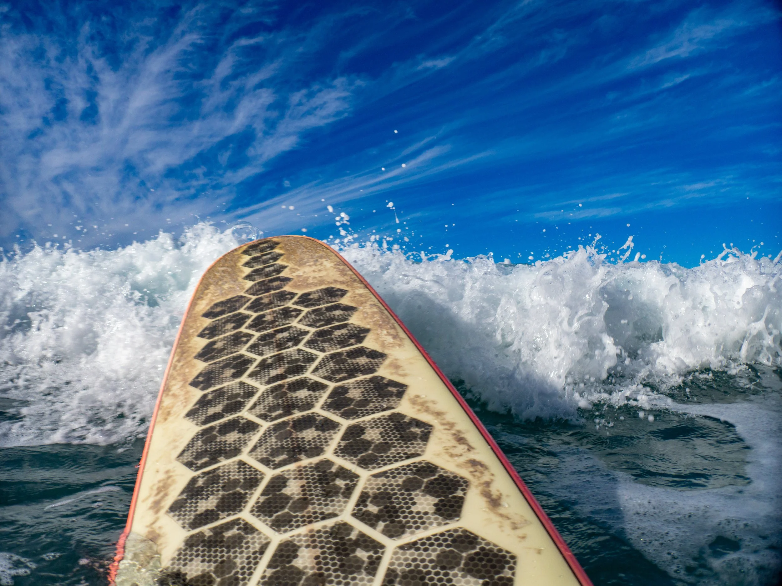 A surfboard with a honeycomb pattern on the bottom, riding a wave in the ocean under a blue sky with scattered clouds.