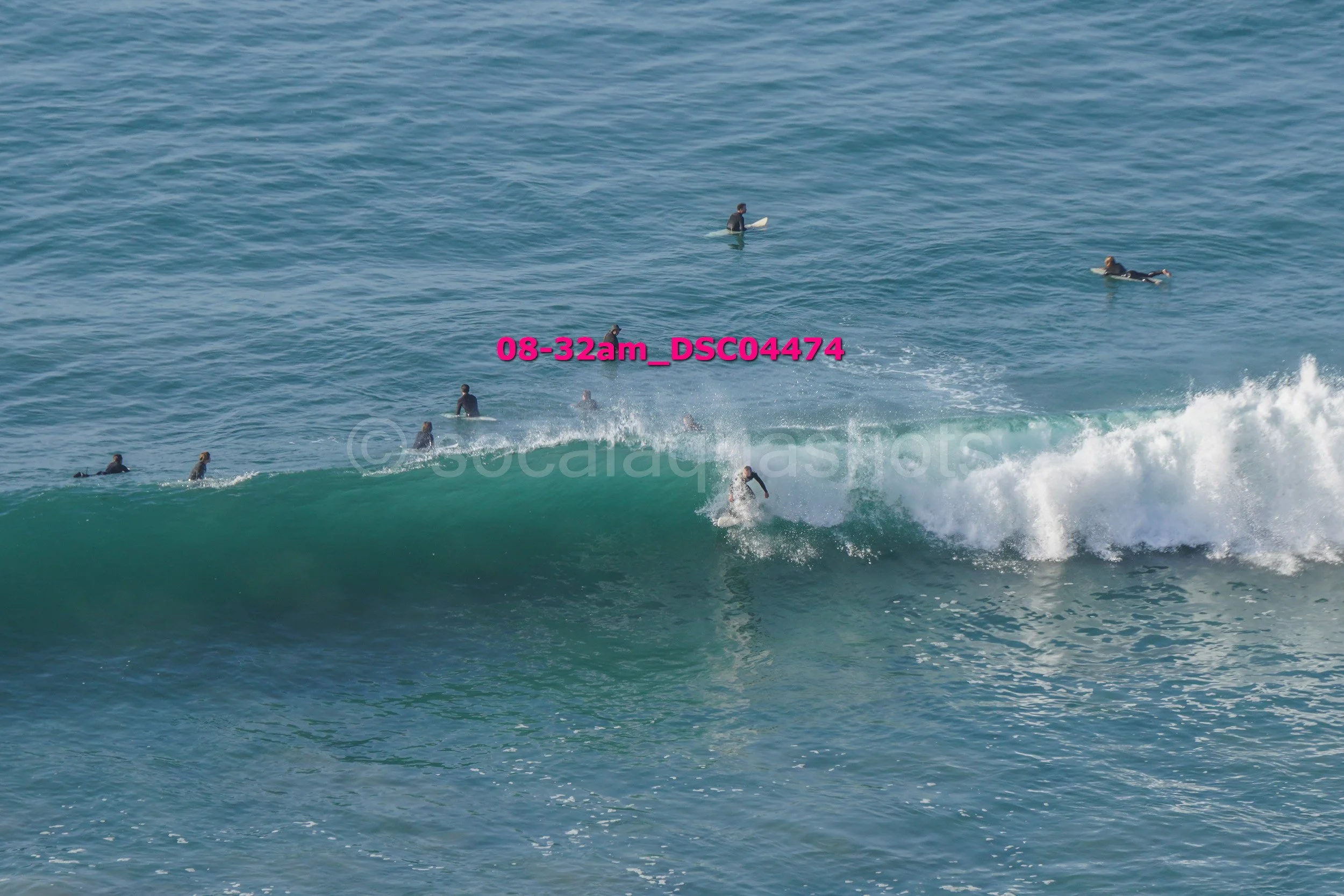 A group of surfers waiting for waves in the ocean, with one surfer riding a wave and others floating on their boards.