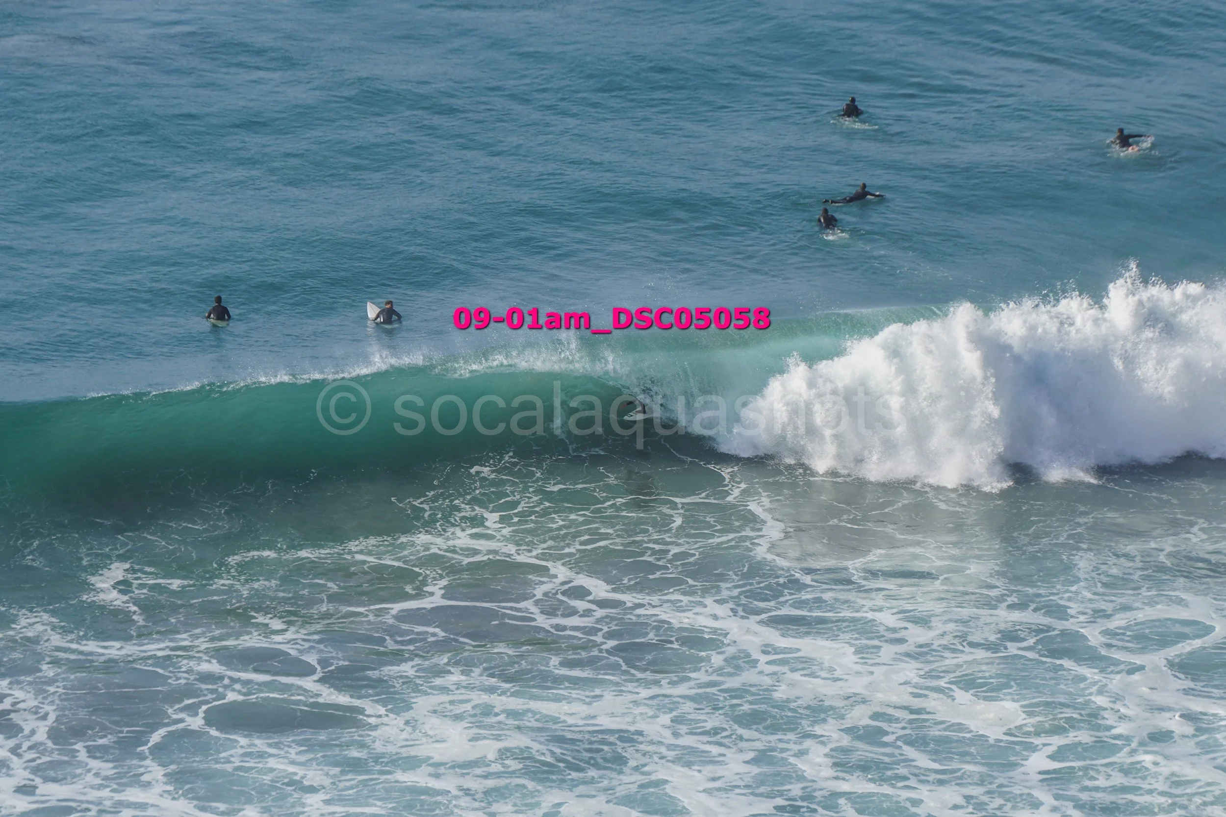 Multiple surfers in wetsuits waiting in the ocean for waves, with one surfer riding a wave.