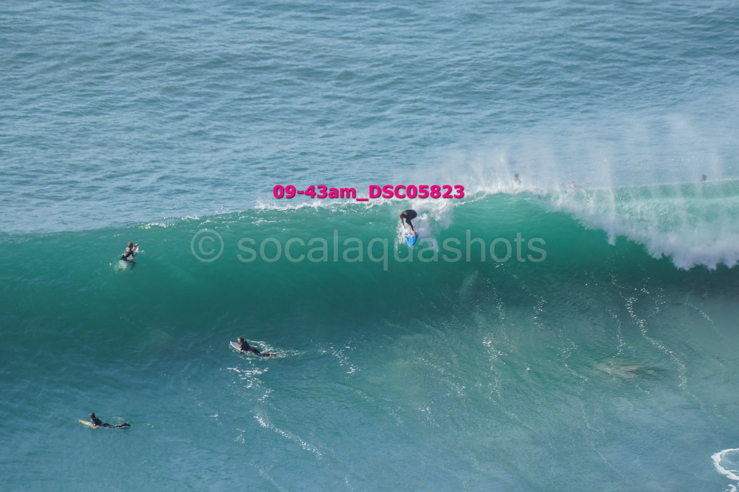A group of surfers riding a large ocean wave, with one surfer performing a maneuver on the wave's lip while others wait in the water.