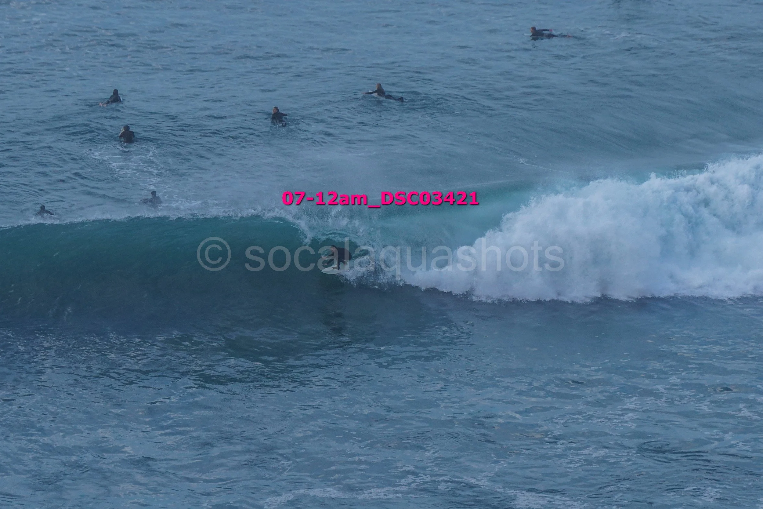 A photograph of a group of surfers in the ocean, with one surfer riding a wave while others wait further out.