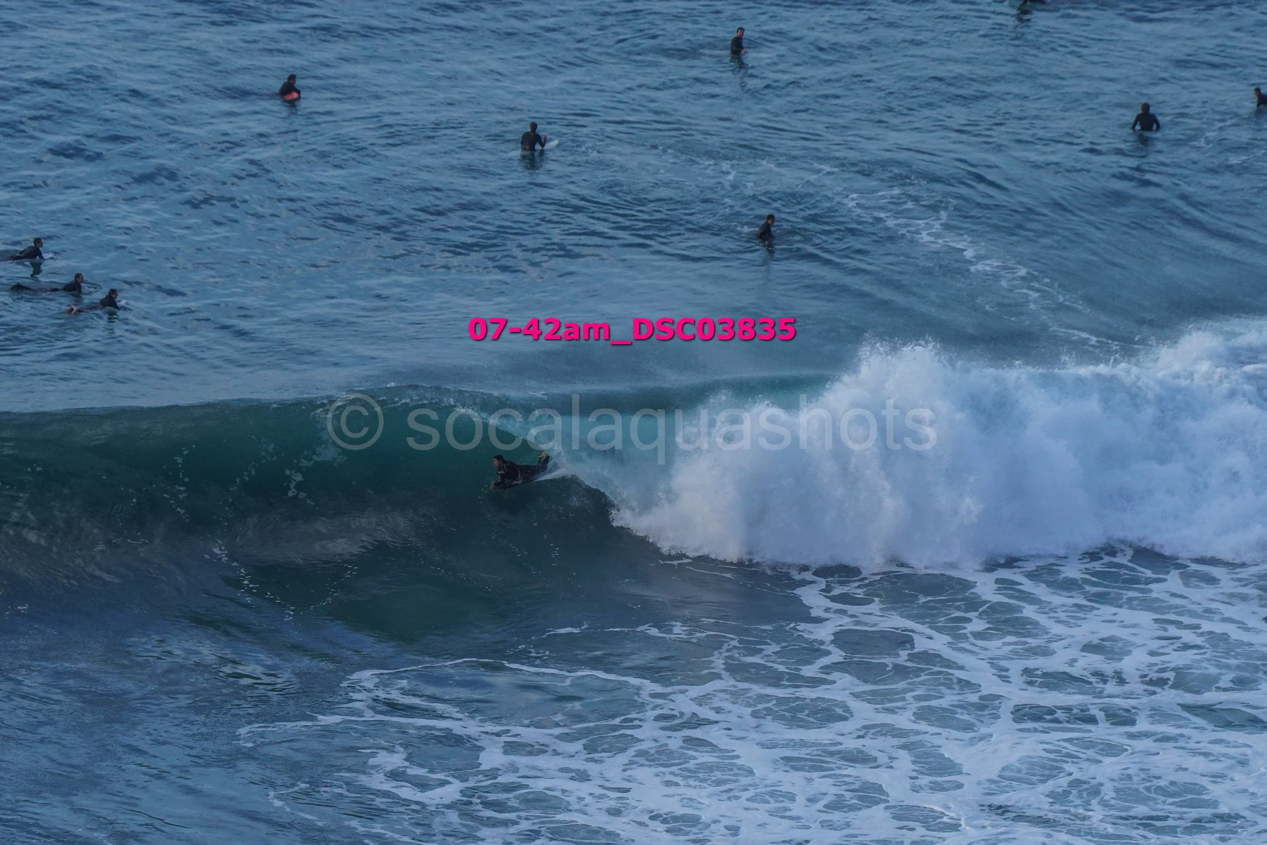 A surfer riding a wave while multiple people in wetsuits are in the water in the background.