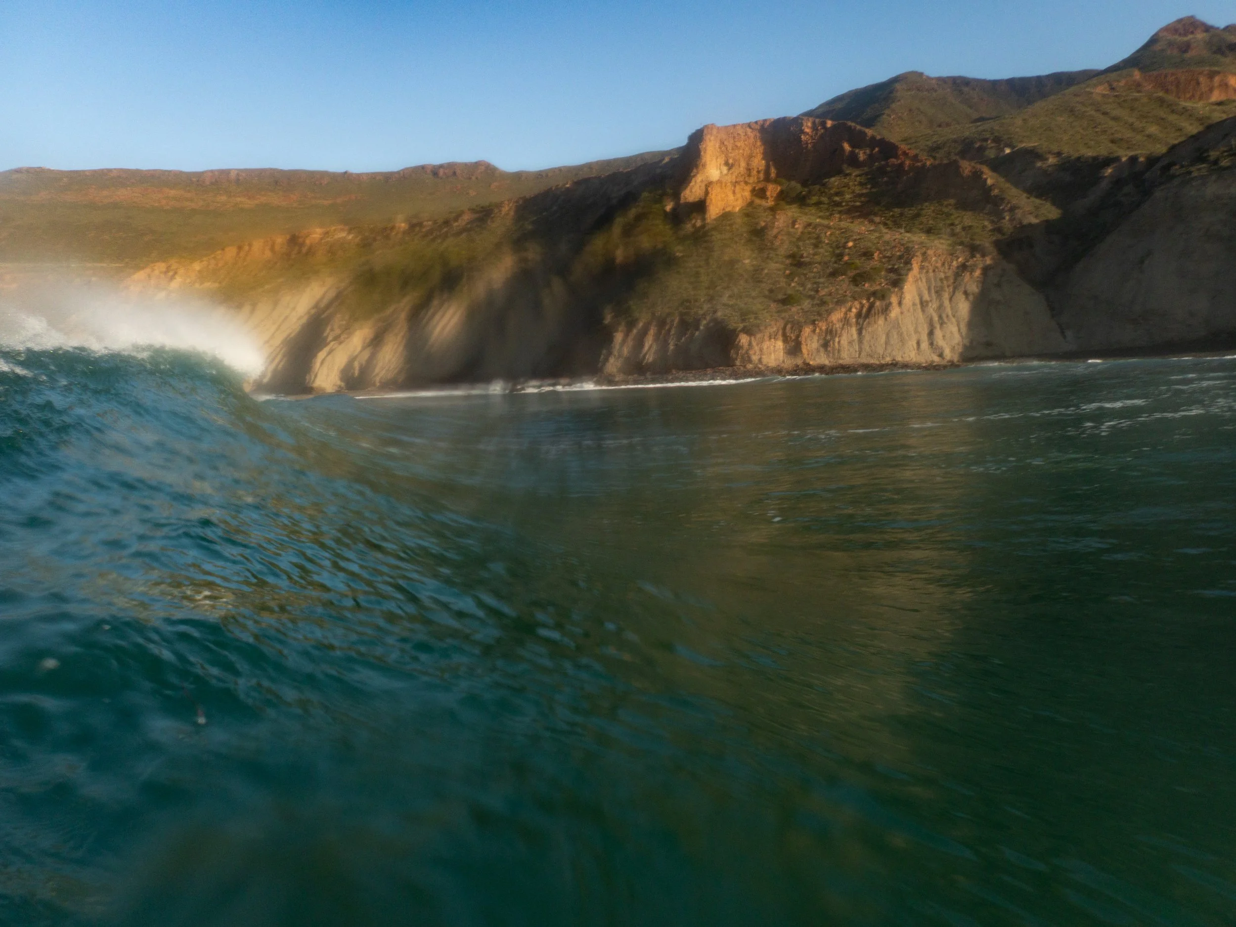 Ocean wave crashing near a rugged coastline with cliffs and hills in the background during sunset.