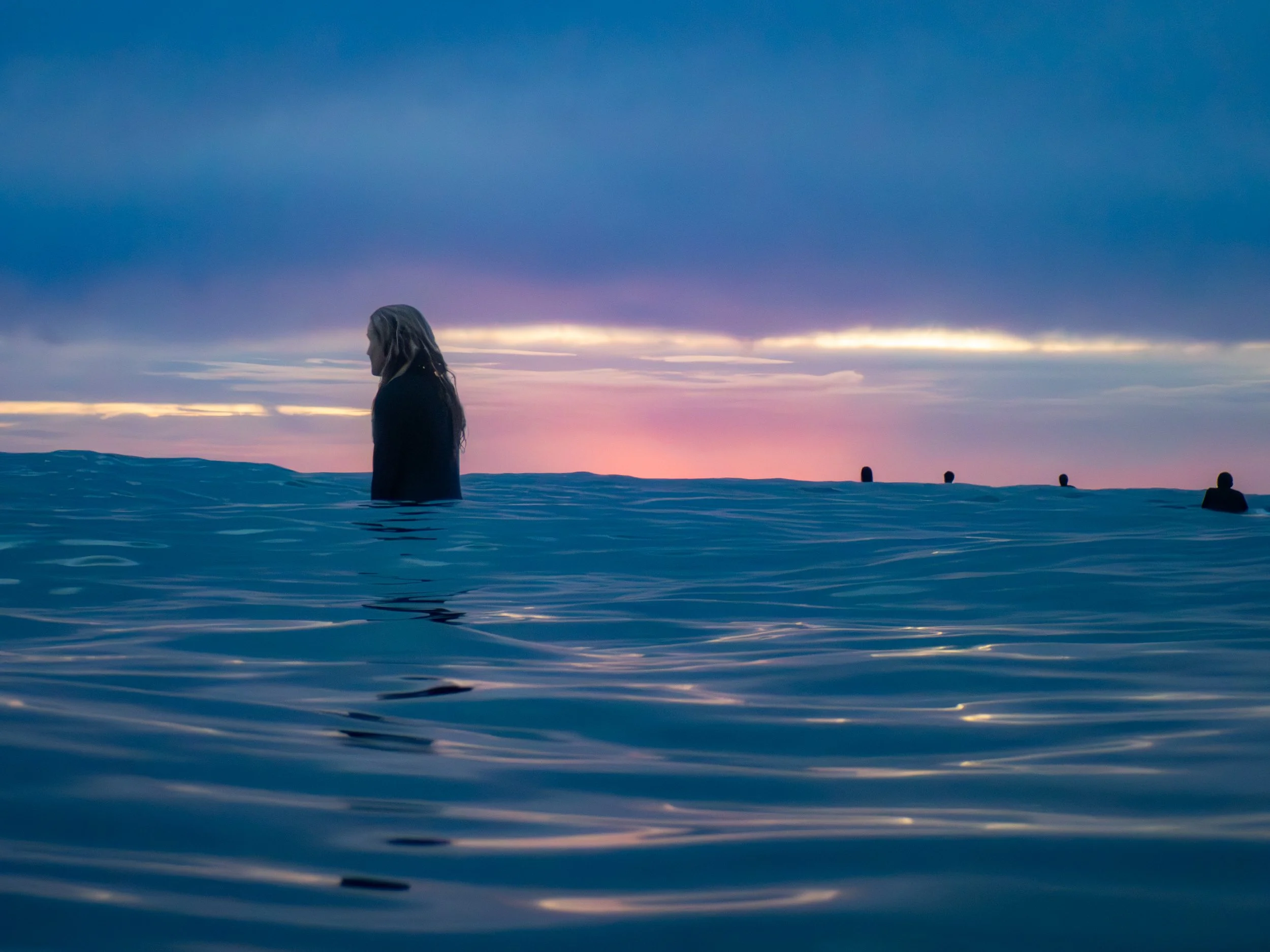 A woman with long hair standing in the ocean during sunset, with a colorful sky and a few other figures in the distance.