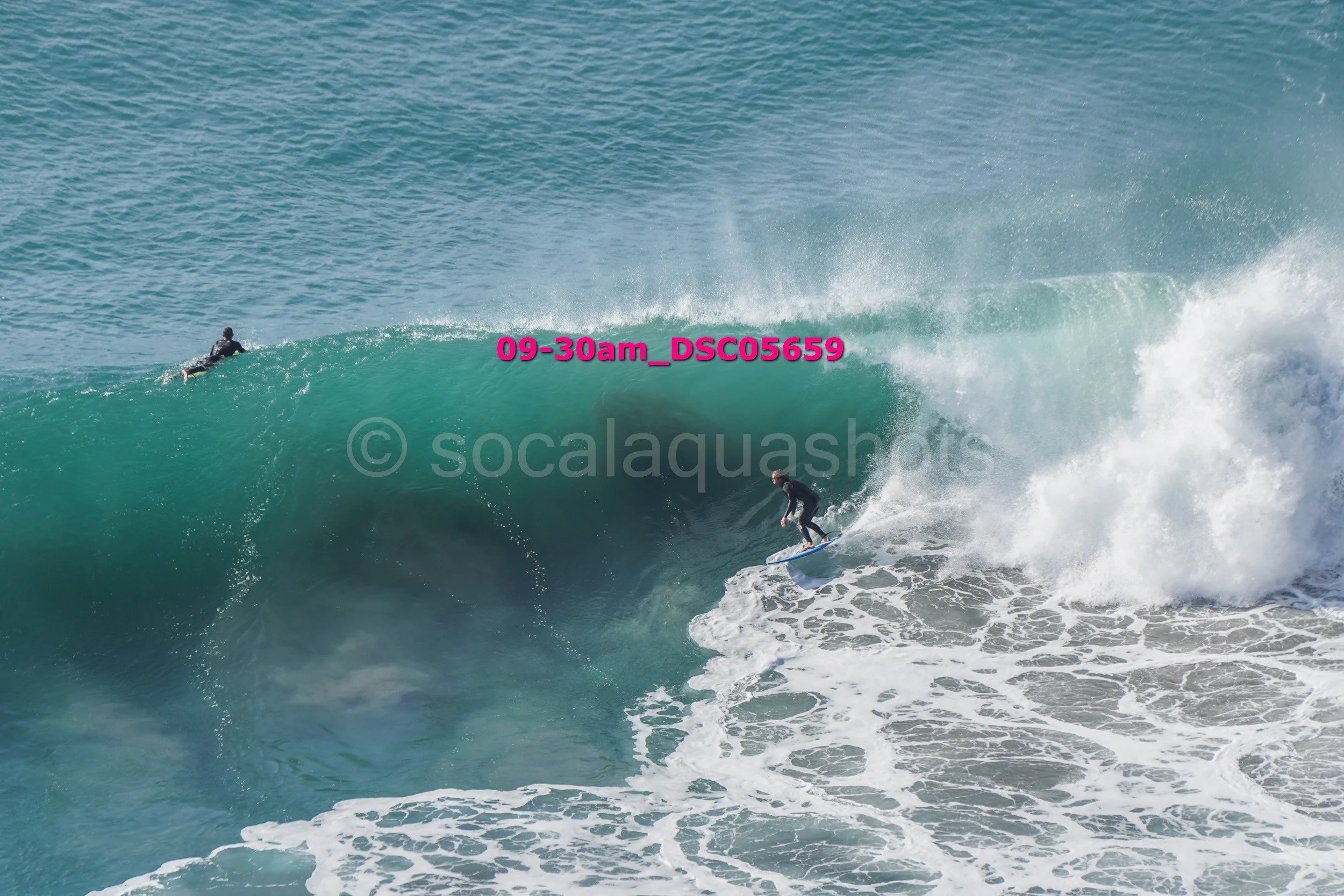 Surfer riding a large green wave, with another surfer in the background on the water, in the ocean with visible white foamy surf.