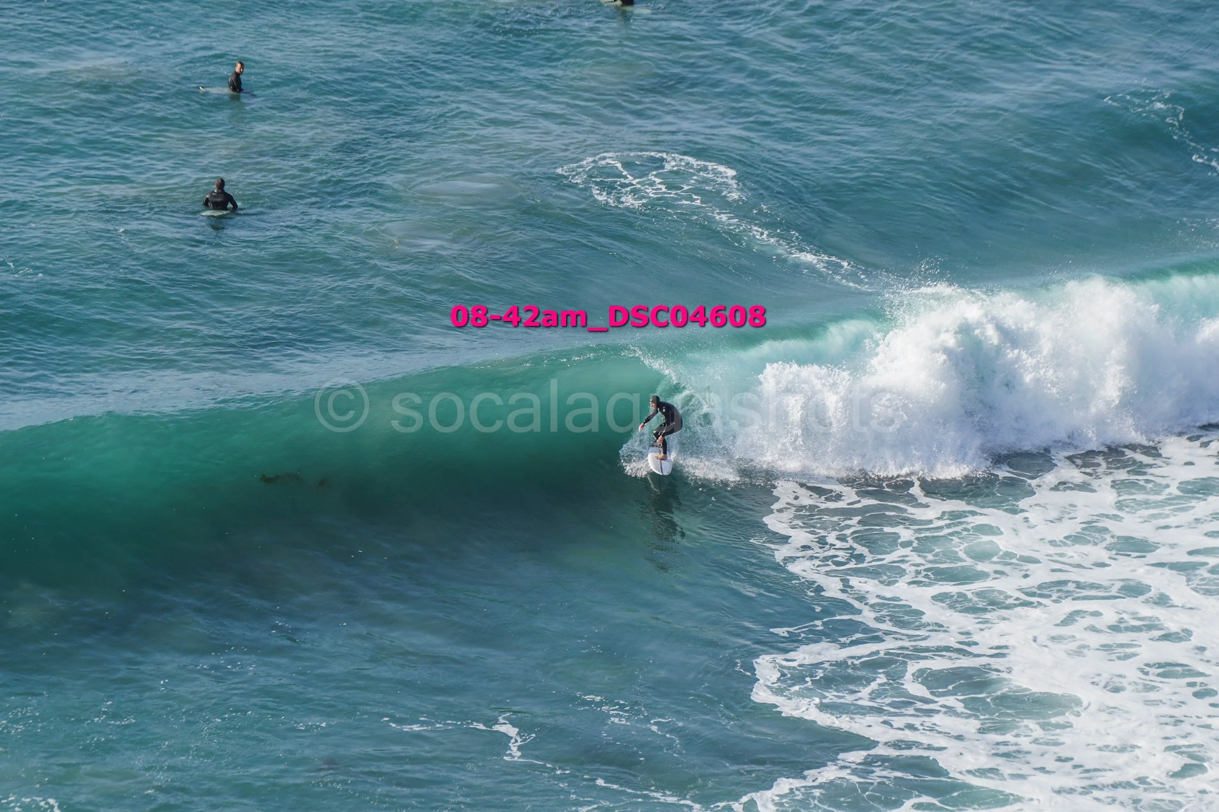 A person surfing on a wave in the ocean, with two other people sitting on surfboards nearby in the water.