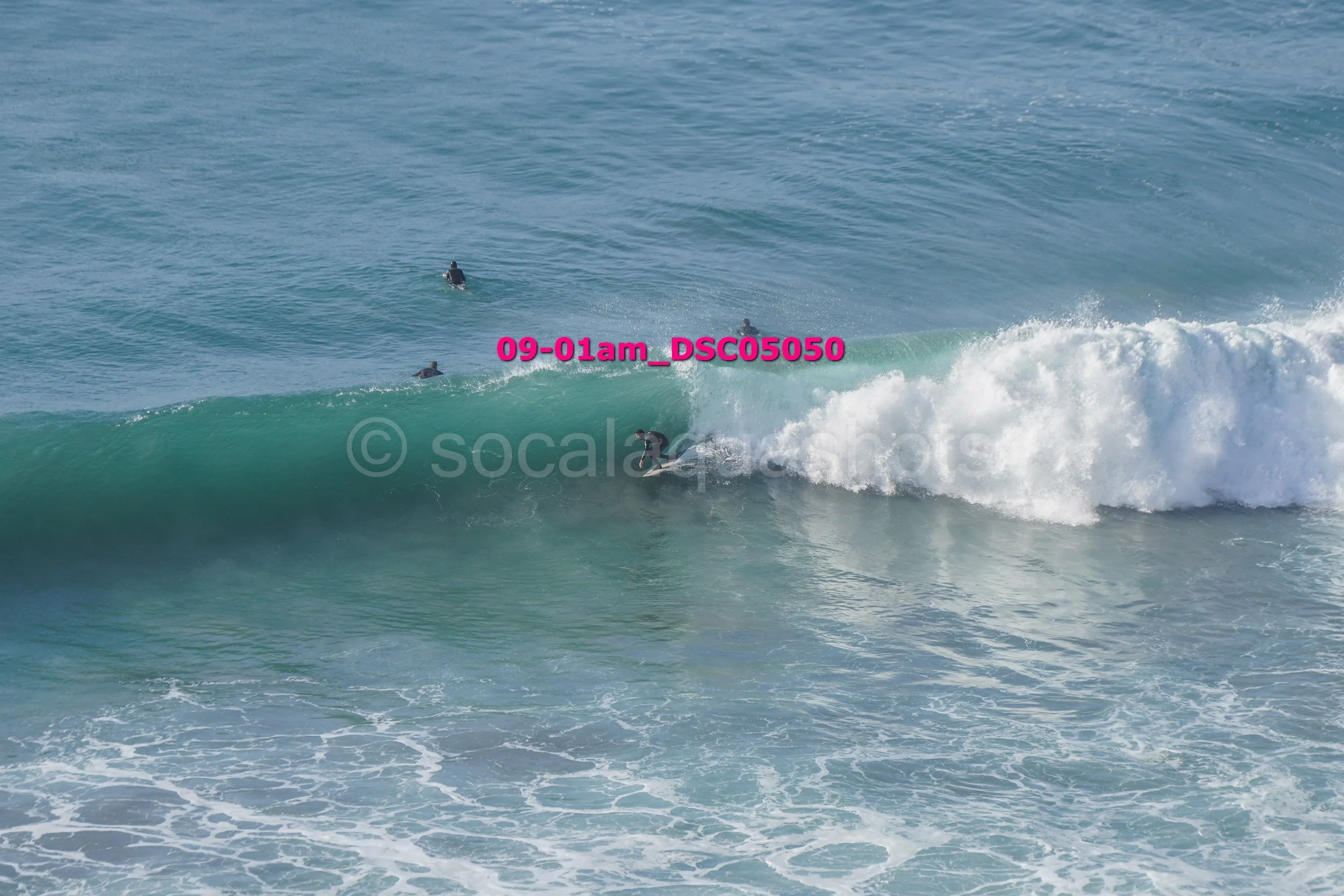 Surfers catching waves in the ocean during daytime, with some waiting and others riding the surf.
