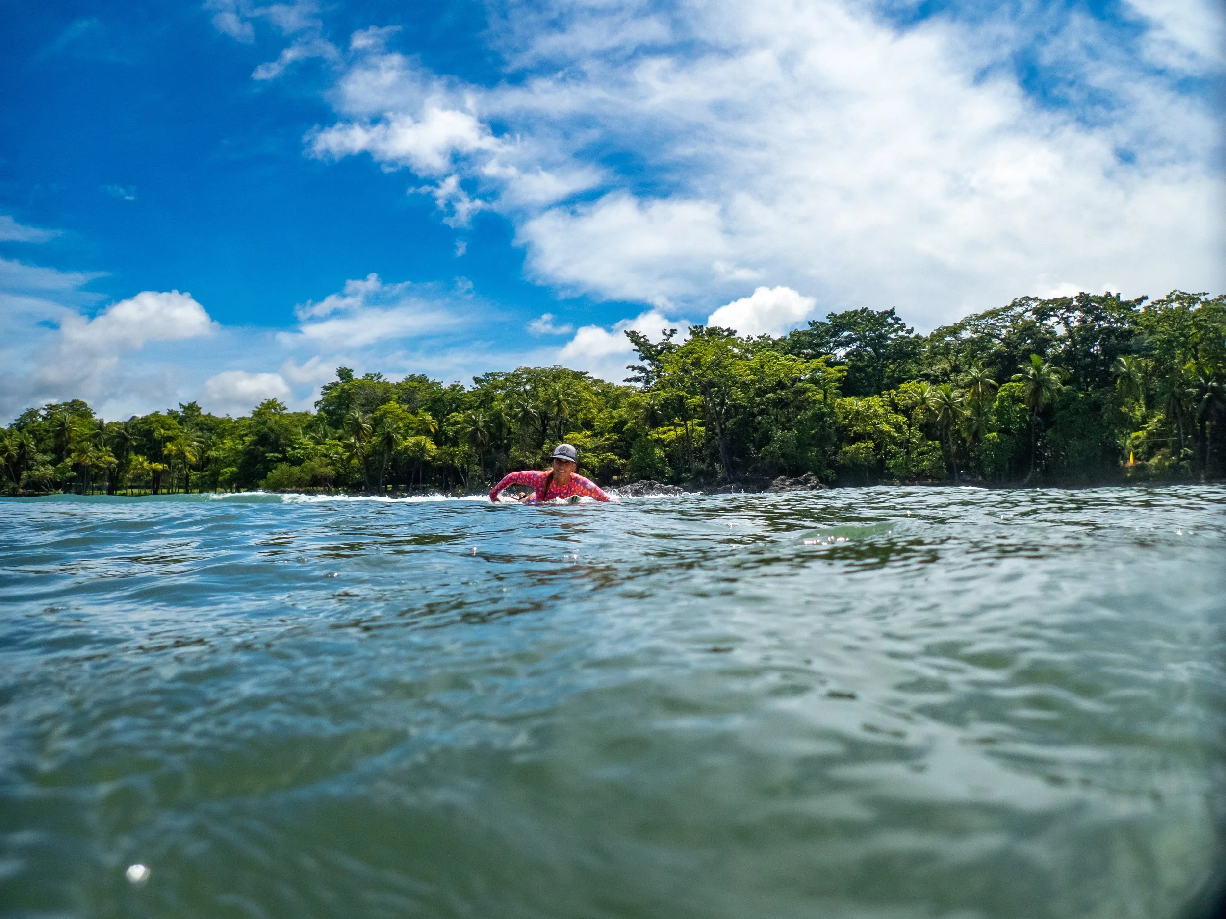 Person in colorful swimsuit swimming in a lake with lush greenery and trees on the bank under a partly cloudy sky.