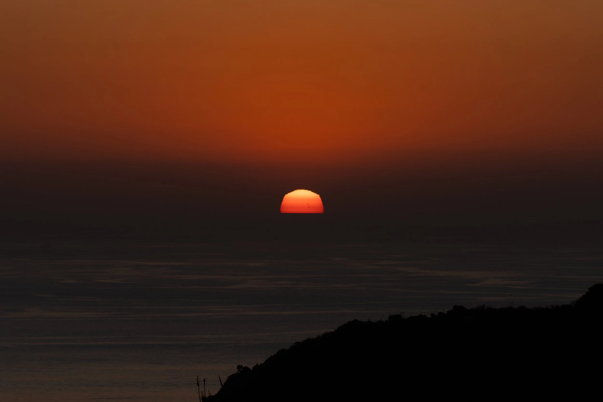 Sunset over the ocean with orange and dark sky, silhouette of a hill in the foreground.
