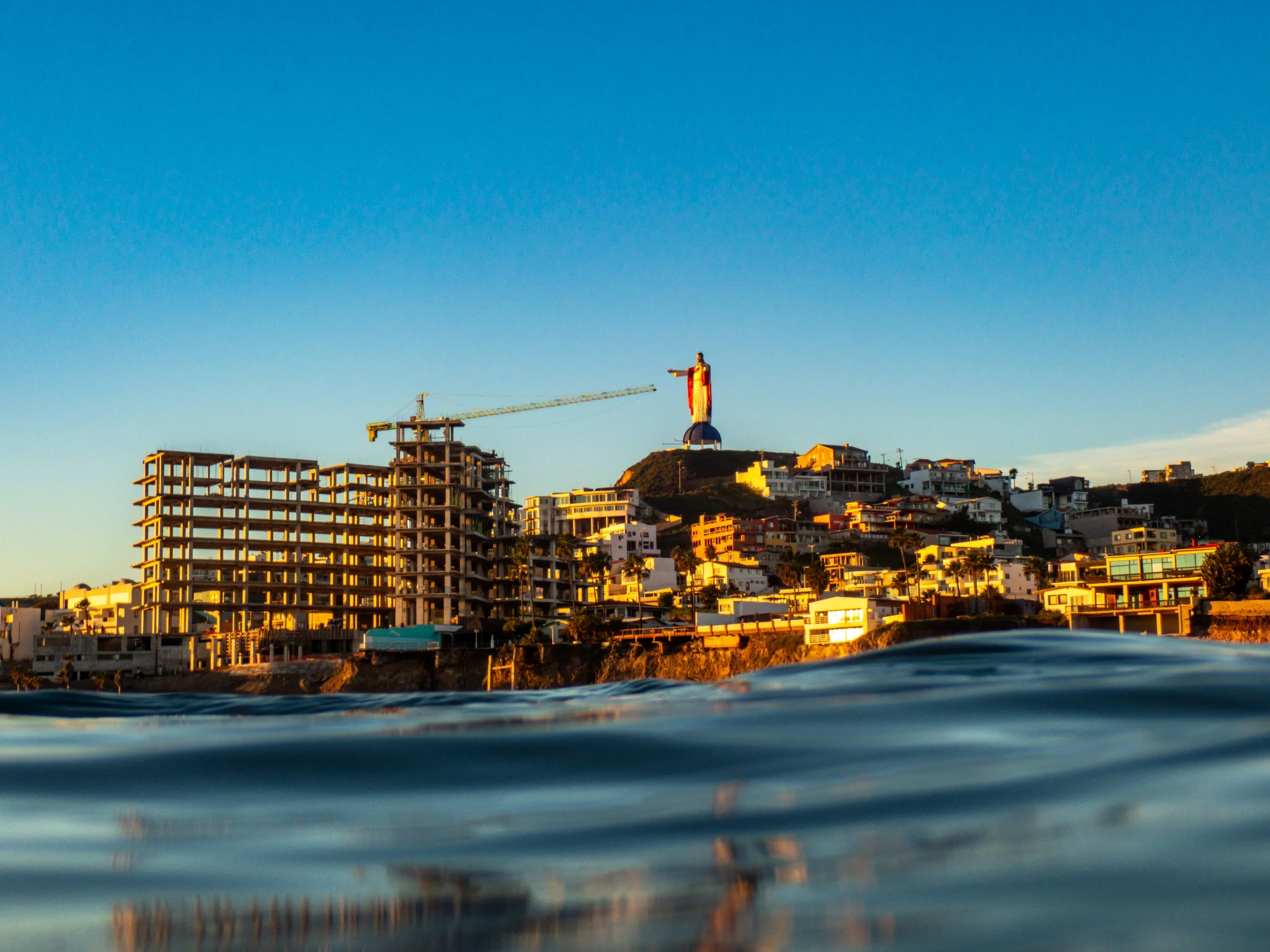 San Diego coastline with the Christ the King statue on a hill in the background, buildings and construction sites along the shore, and water in the foreground, under a clear blue sky during sunset.