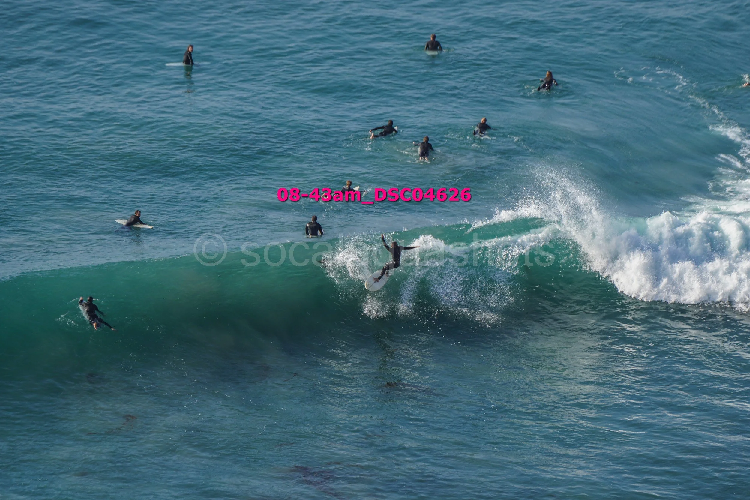 A group of people surfing in the ocean, with one person riding a wave and others waiting on surfboards or swimming in the water.