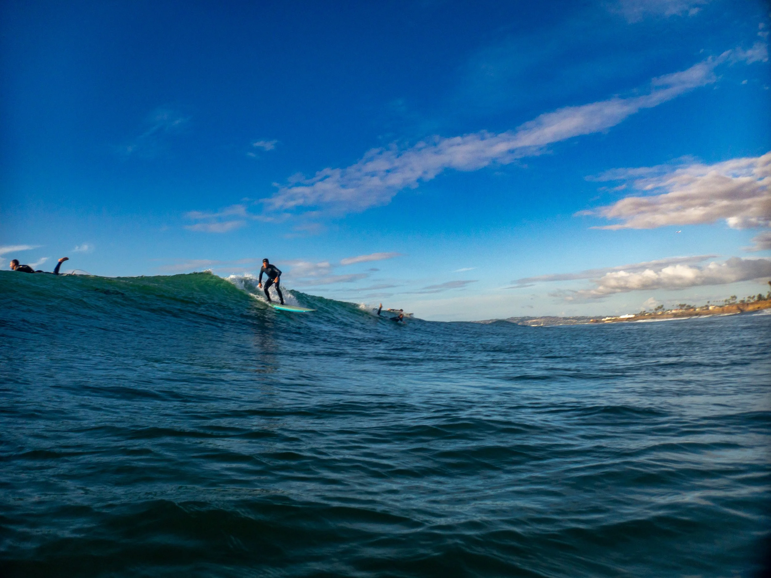 Surfer riding a wave in the ocean under a partly cloudy sky with other surfers in the water and land in the distance.