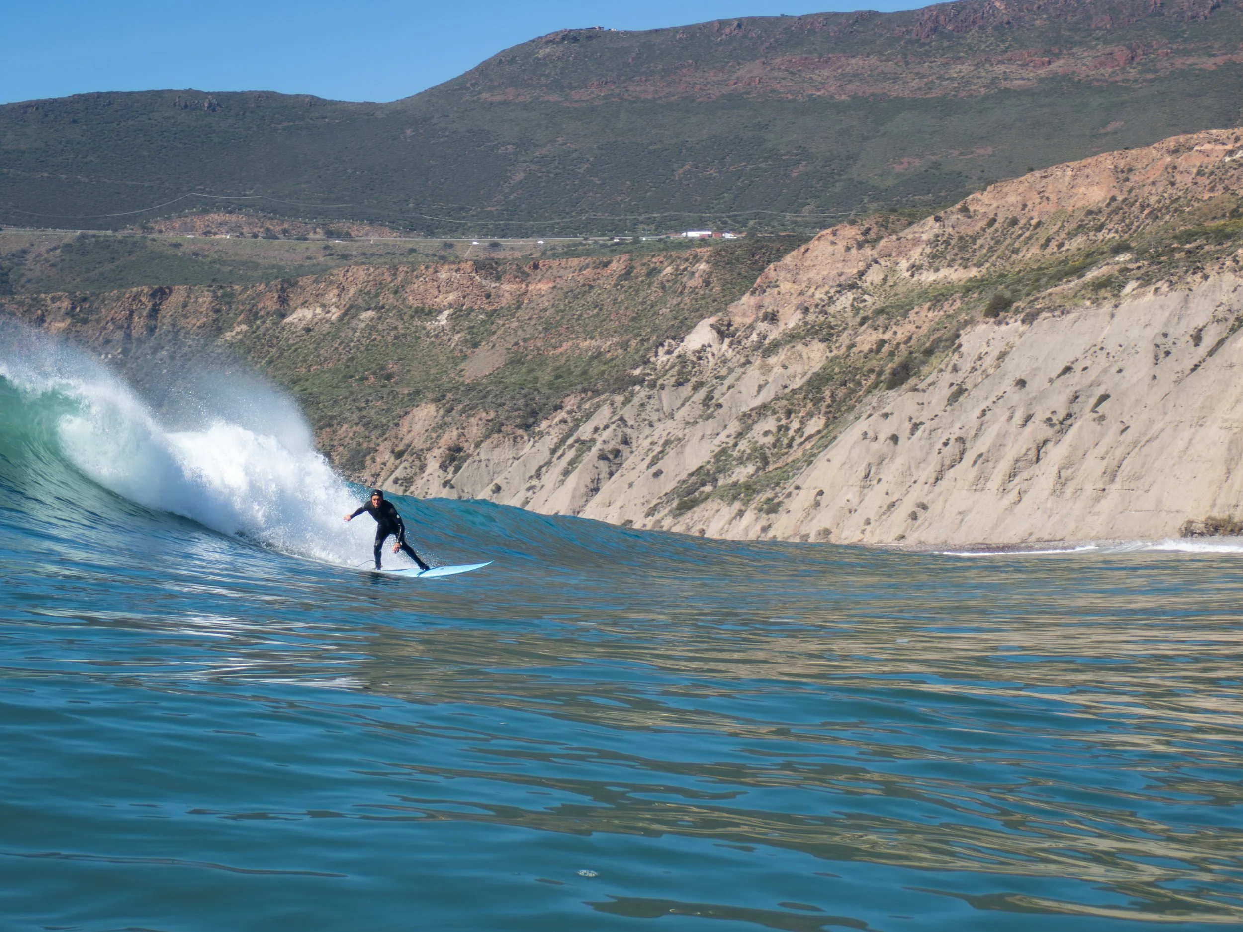 A person surfing on a wave near a rocky coastline and hillside under a clear blue sky.