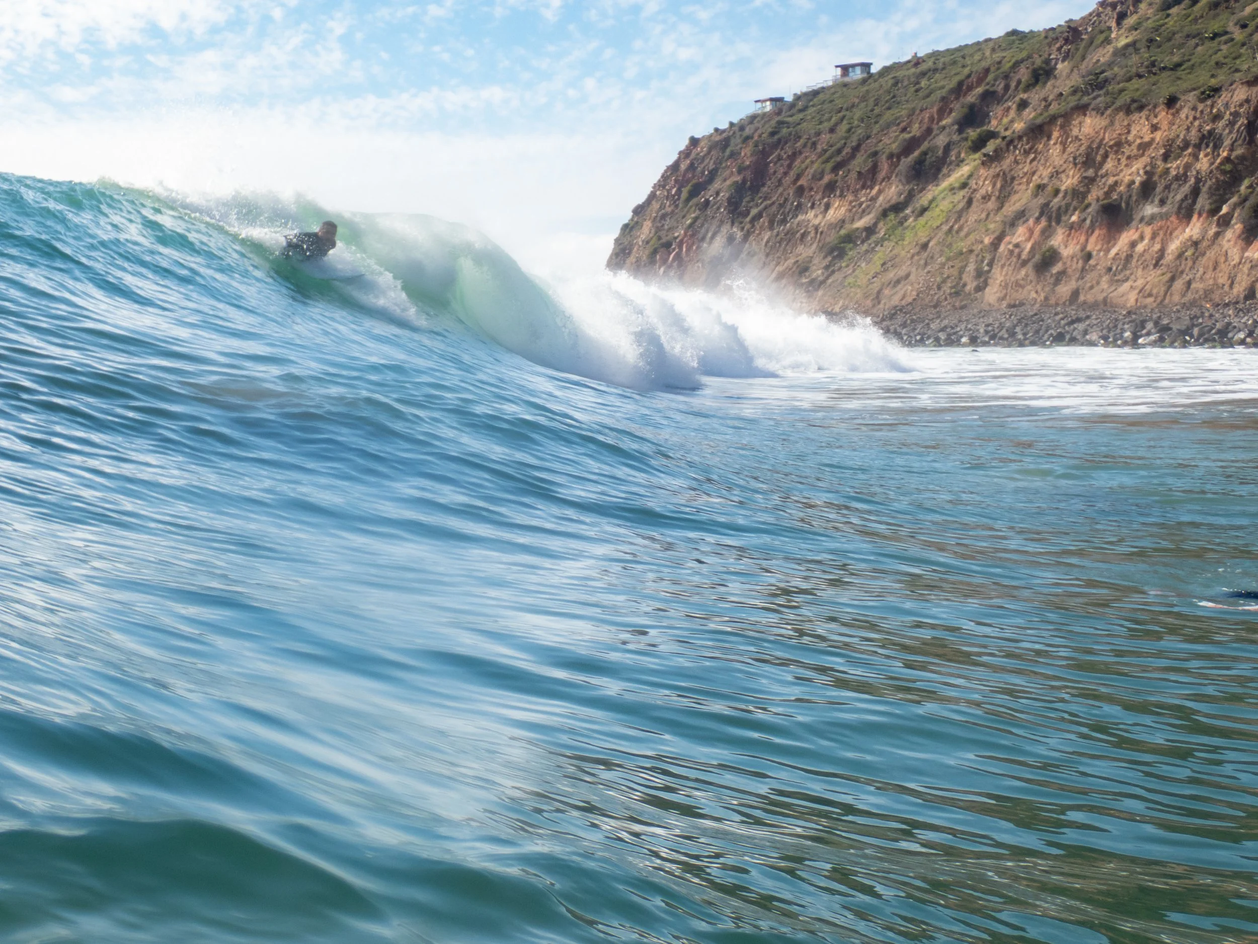 A person surfing on a wave near a rocky coastal shoreline with a hill in the background.