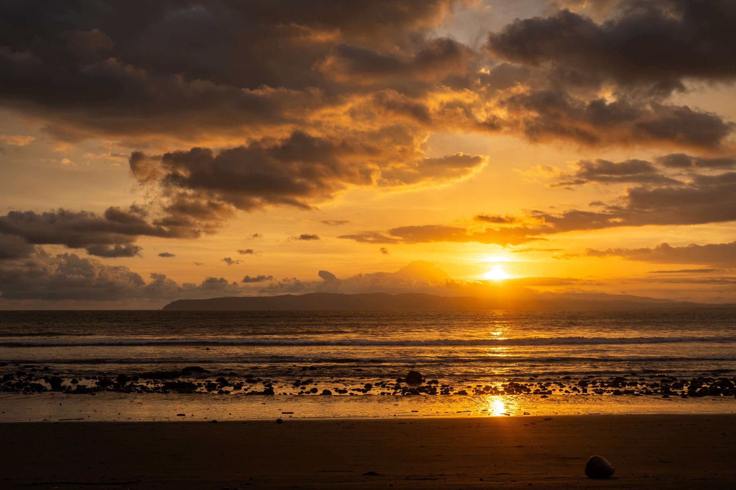 A sunset over the ocean with dark clouds in the sky, a distant landmass on the horizon, and rocks on the beach reflecting the golden glow.