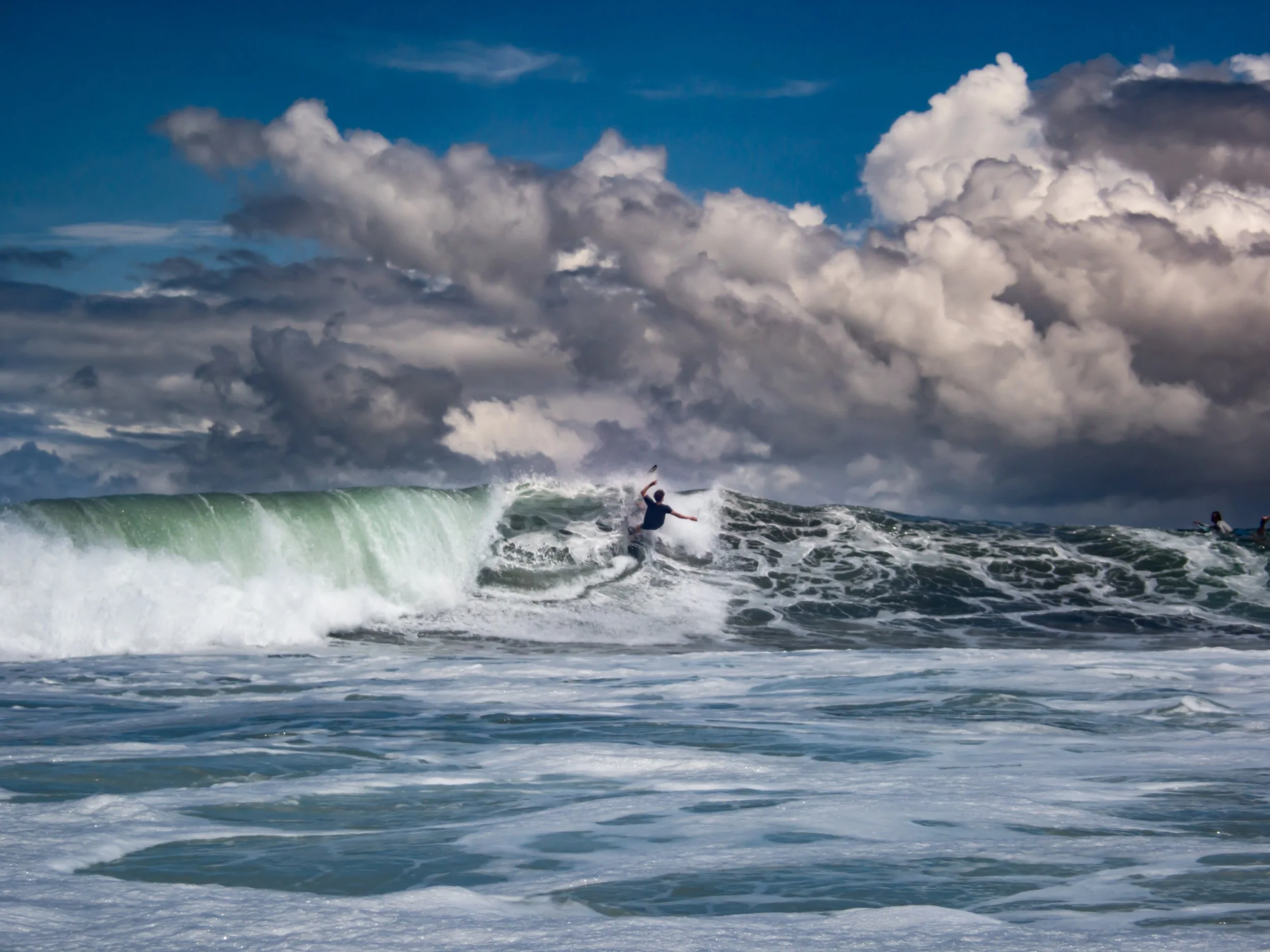 Surfer riding a large ocean wave under dramatic cloudy sky.