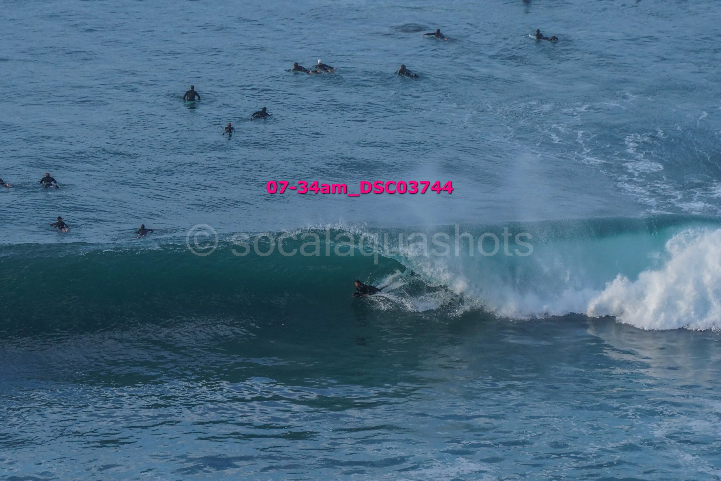 A person surfing on a large wave with many people in the water watching from above.