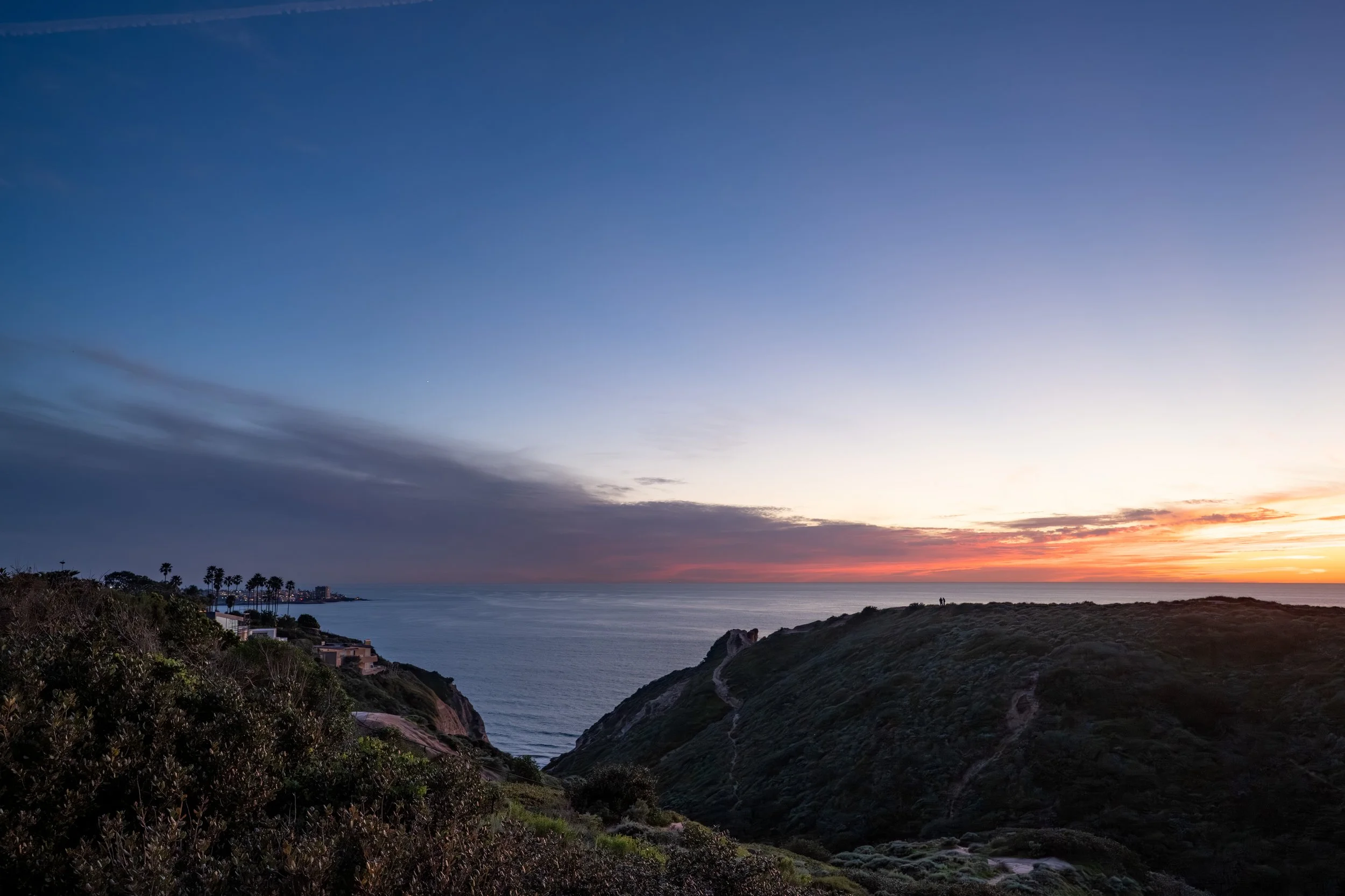 Seaside view during sunset with a sky transitioning from blue to pink, featuring cliffs covered in green vegetation and a few houses along the coast.