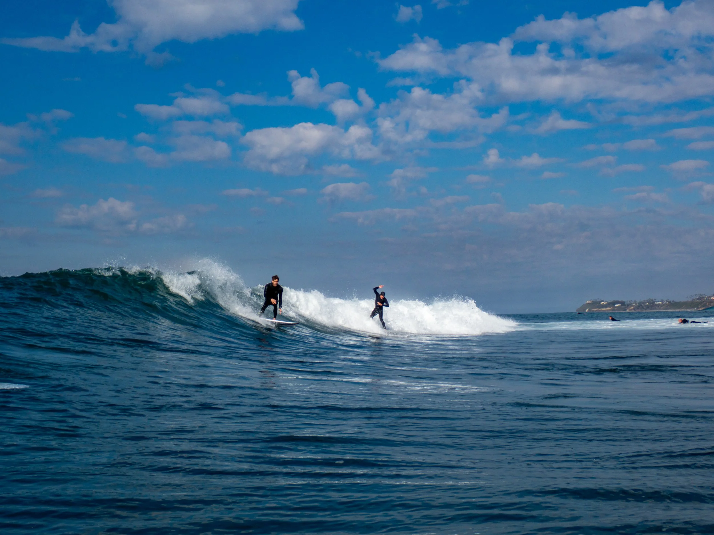 Two surfers riding a wave in the ocean under a partly cloudy sky, with other surfers visible in the distance.
