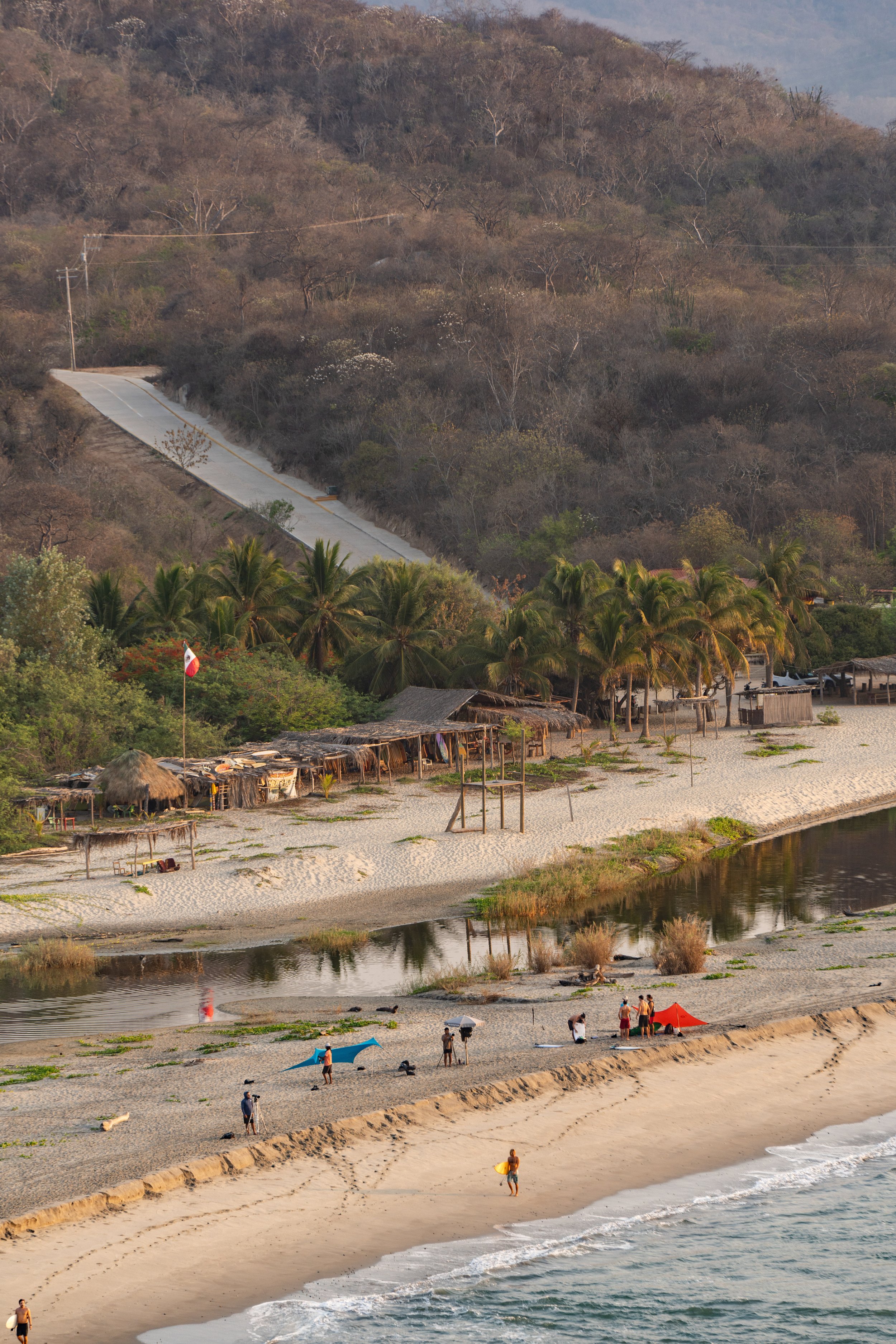 A scenic view of a sandy beach with people under colorful umbrellas, calm water, palm trees, huts, and a hillside with dry trees in the background.