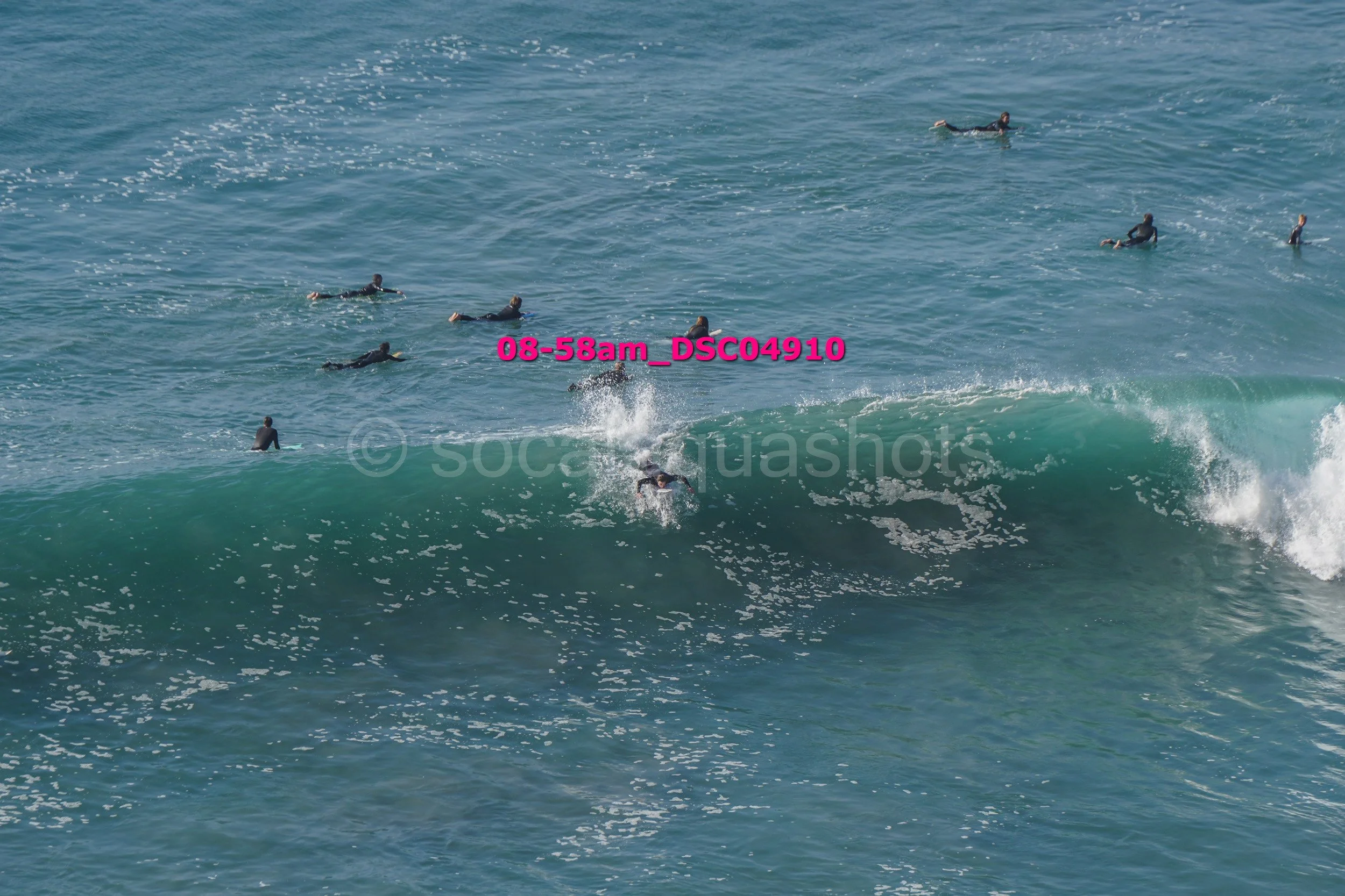 Surfers in wetsuits riding and waiting for waves in the ocean, some on surfboards and some swimming.