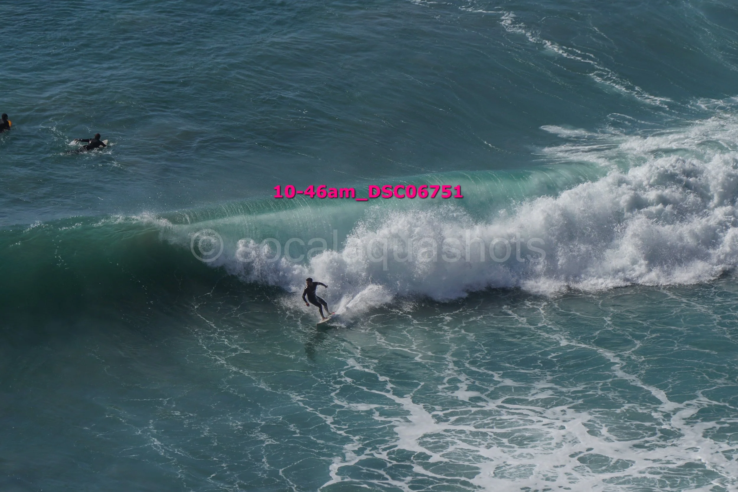 A person surfing on a wave at the beach. There are three other surfers in the water, and the sky is clear.