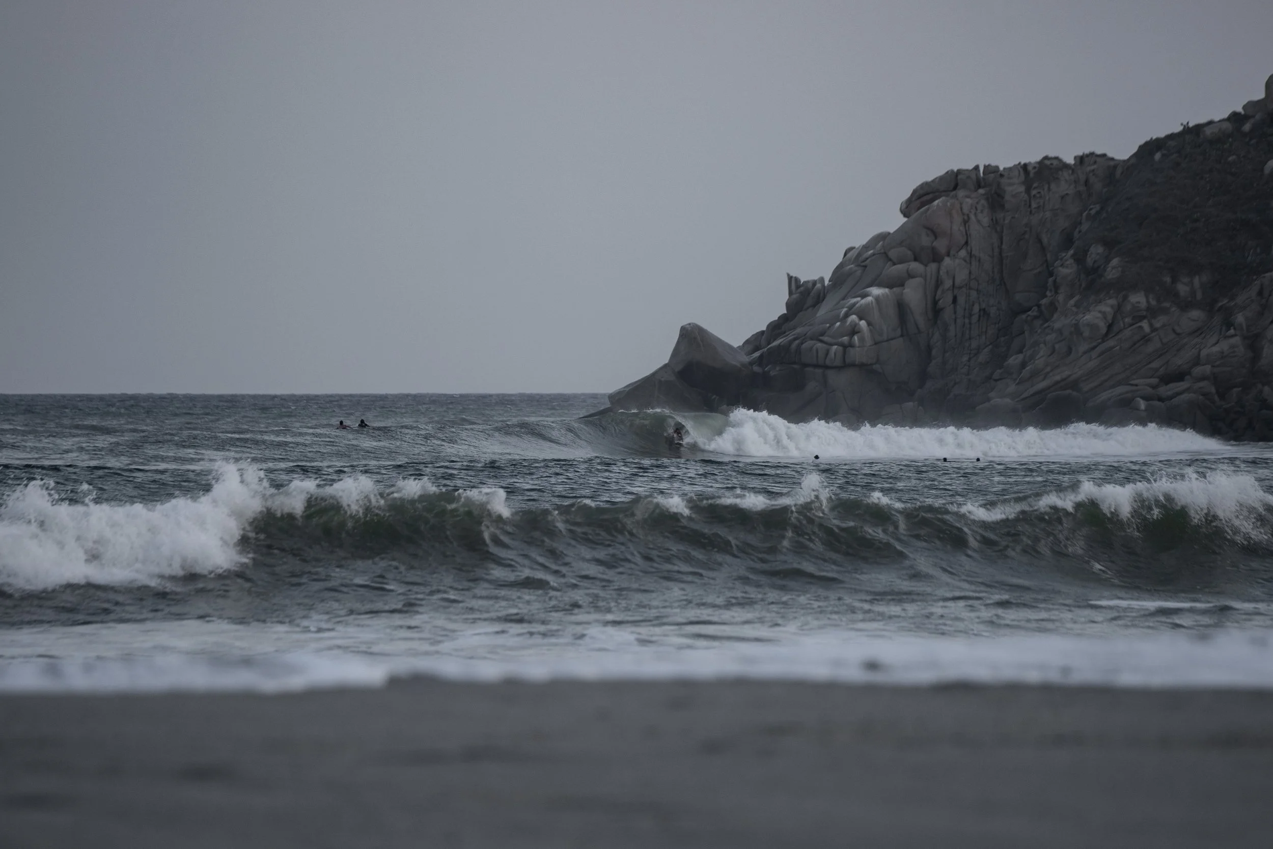 Ocean waves crashing near a rocky cliff on a cloudy day, with some surfers in the water.