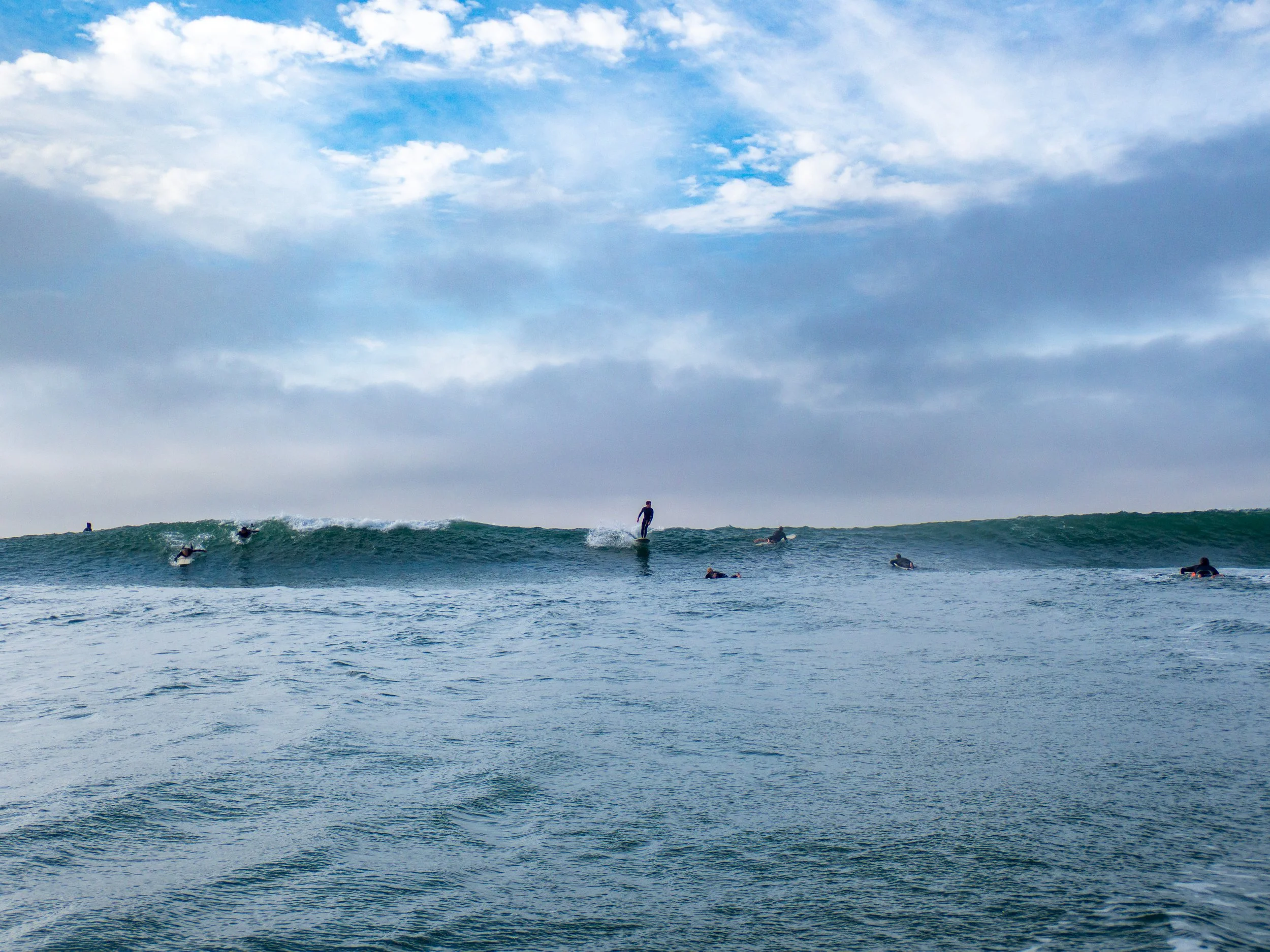 Several surfers in wetsuits surfing and floating on the ocean with a breaking wave, under a partly cloudy sky.