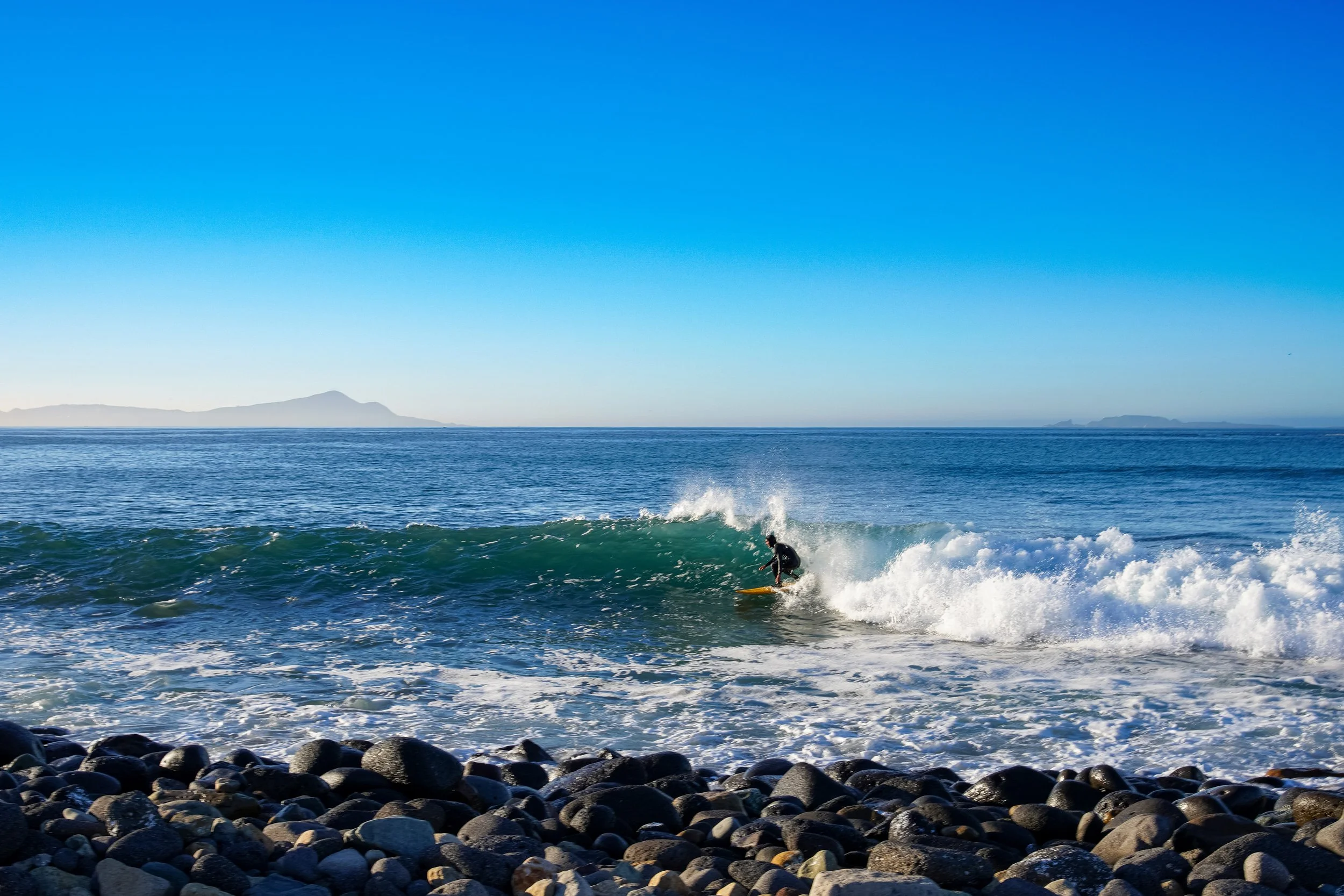 A person surfing on a wave near a rocky beach with mountains in the background under a clear blue sky.