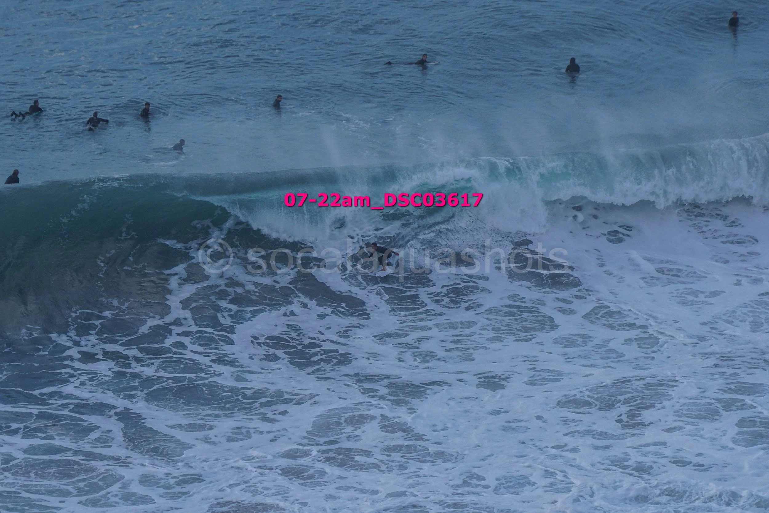 A group of surfers in the ocean, some riding a wave and others in the water, with the sky and horizon in the background.