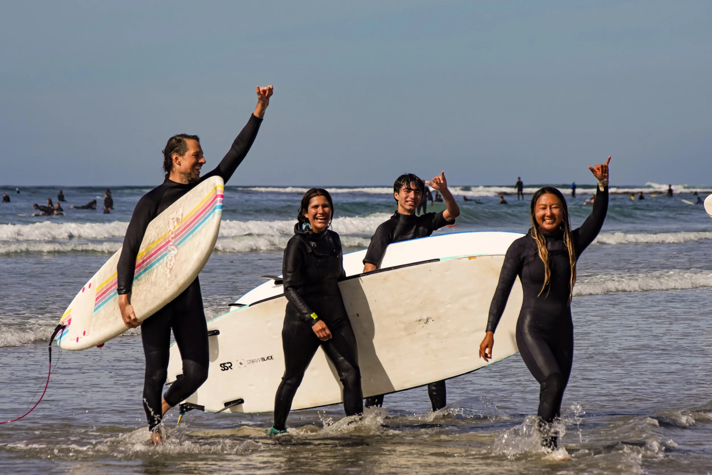 Four surfers in wetsuits walking out of the ocean with surfboards, smiling and celebrating after surfing on a sunny day.