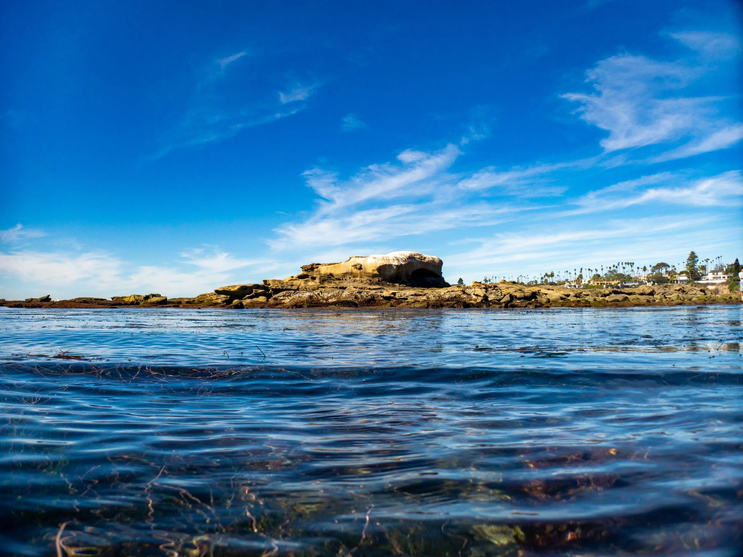 View of rocky shoreline and ocean with clear blue sky and wispy clouds in the background.