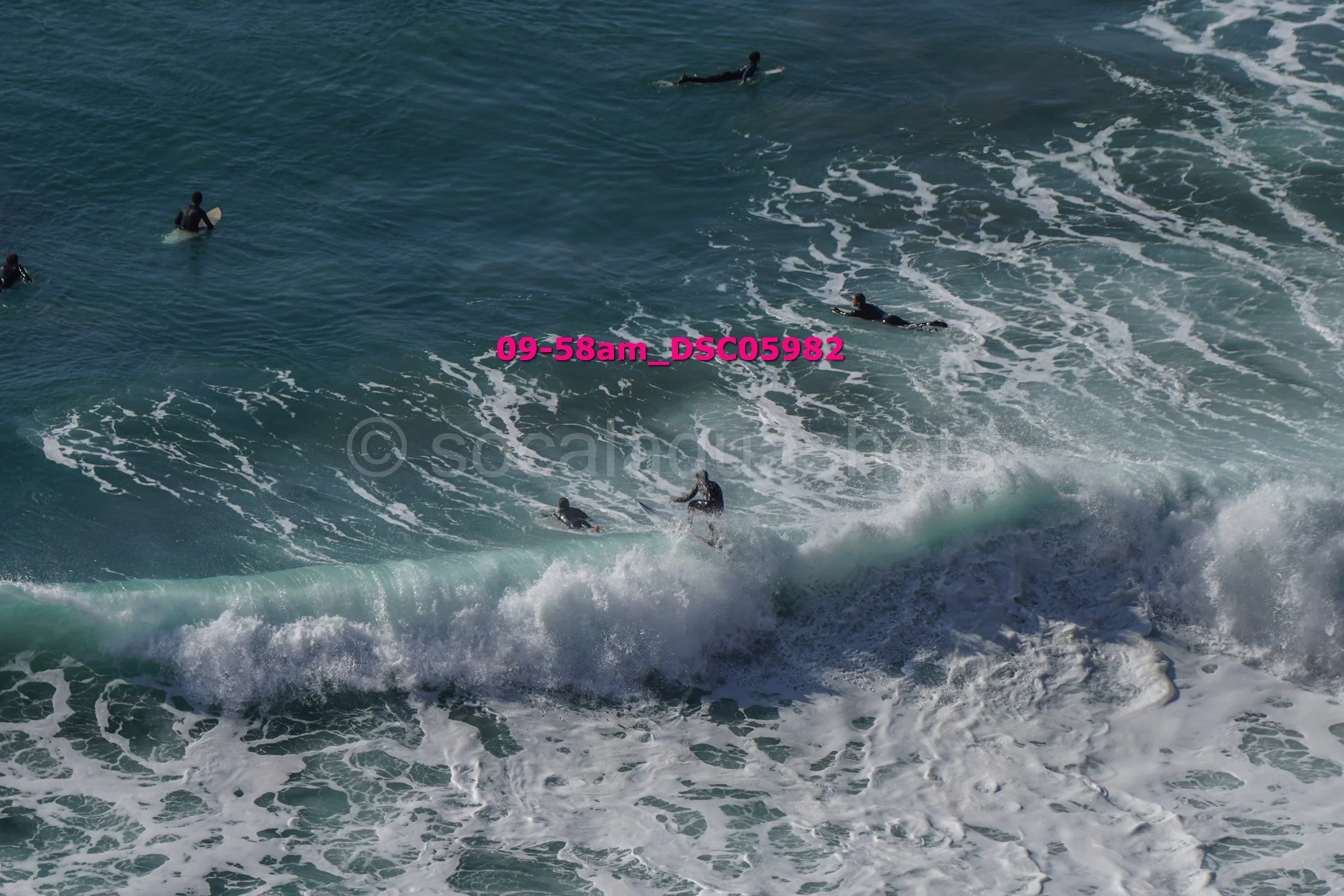 Group of surfers in the ocean waiting for waves, with some riding and others paddling, in a blue sea with white foam.