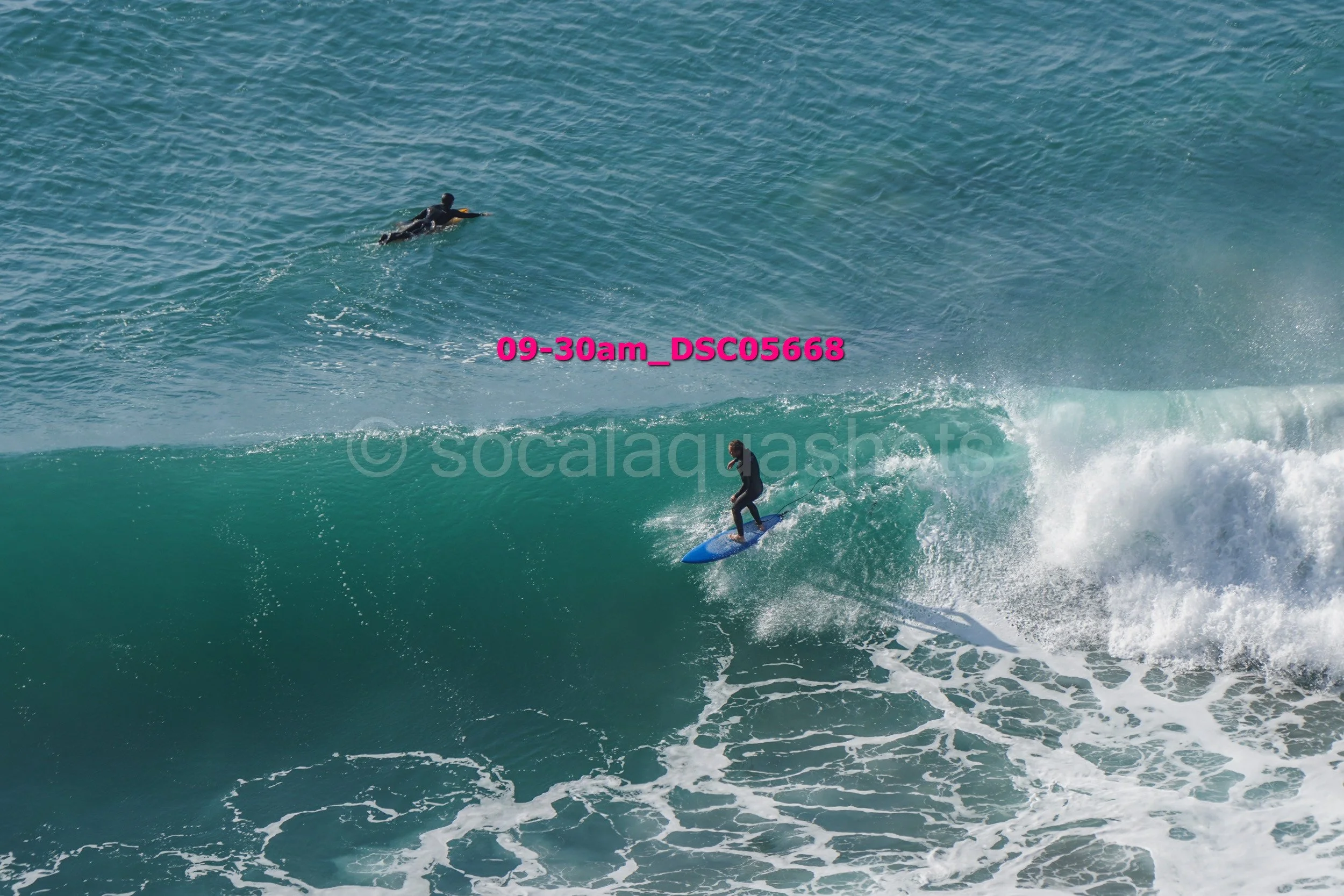 A person surfing on a wave while facing the water, with another person swimming or surfing farther in the distance.