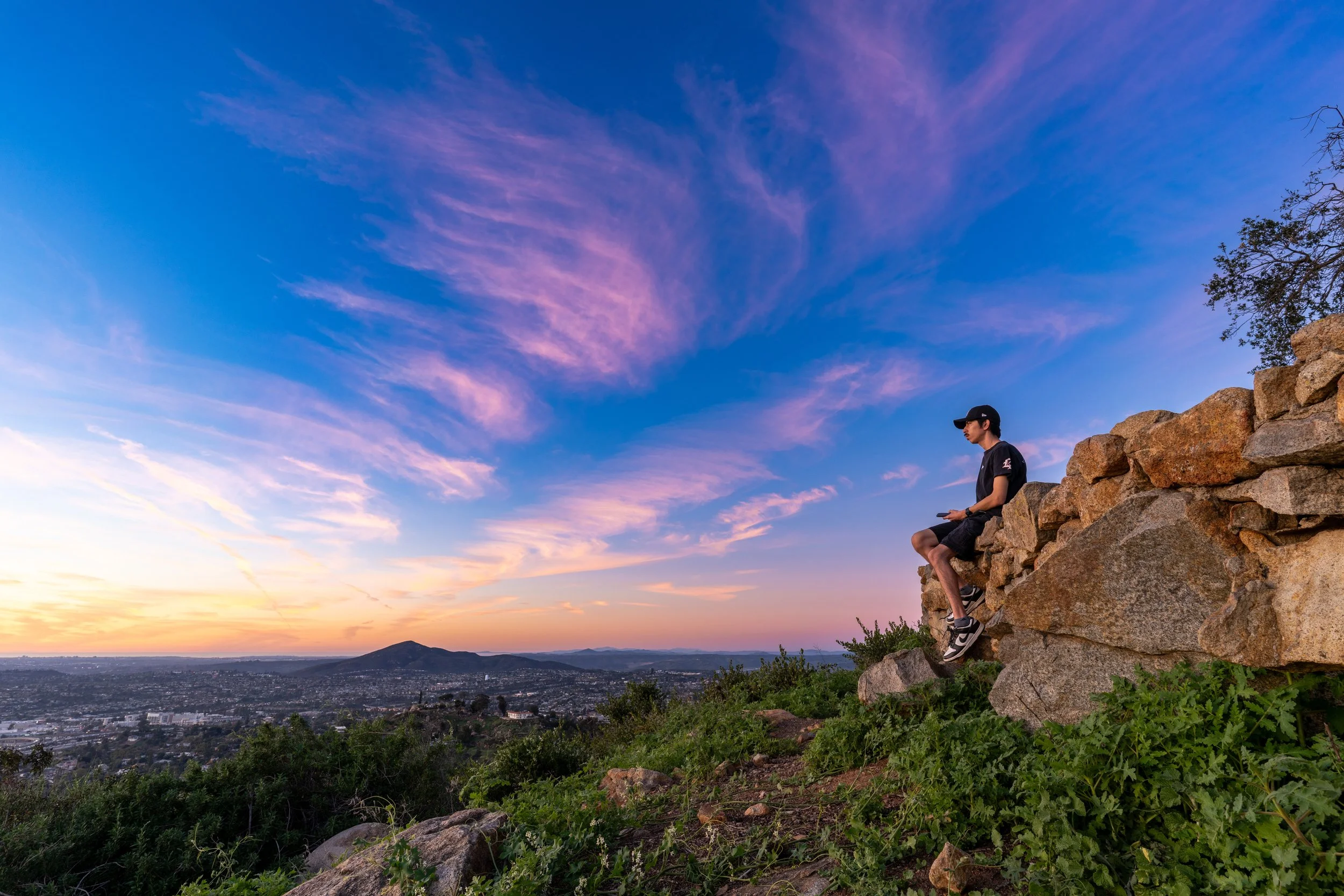 A young man sitting on a rocky cliff edge at sunset, overlooking a city with hills in the distance, under a colorful sky with pink and purple clouds.