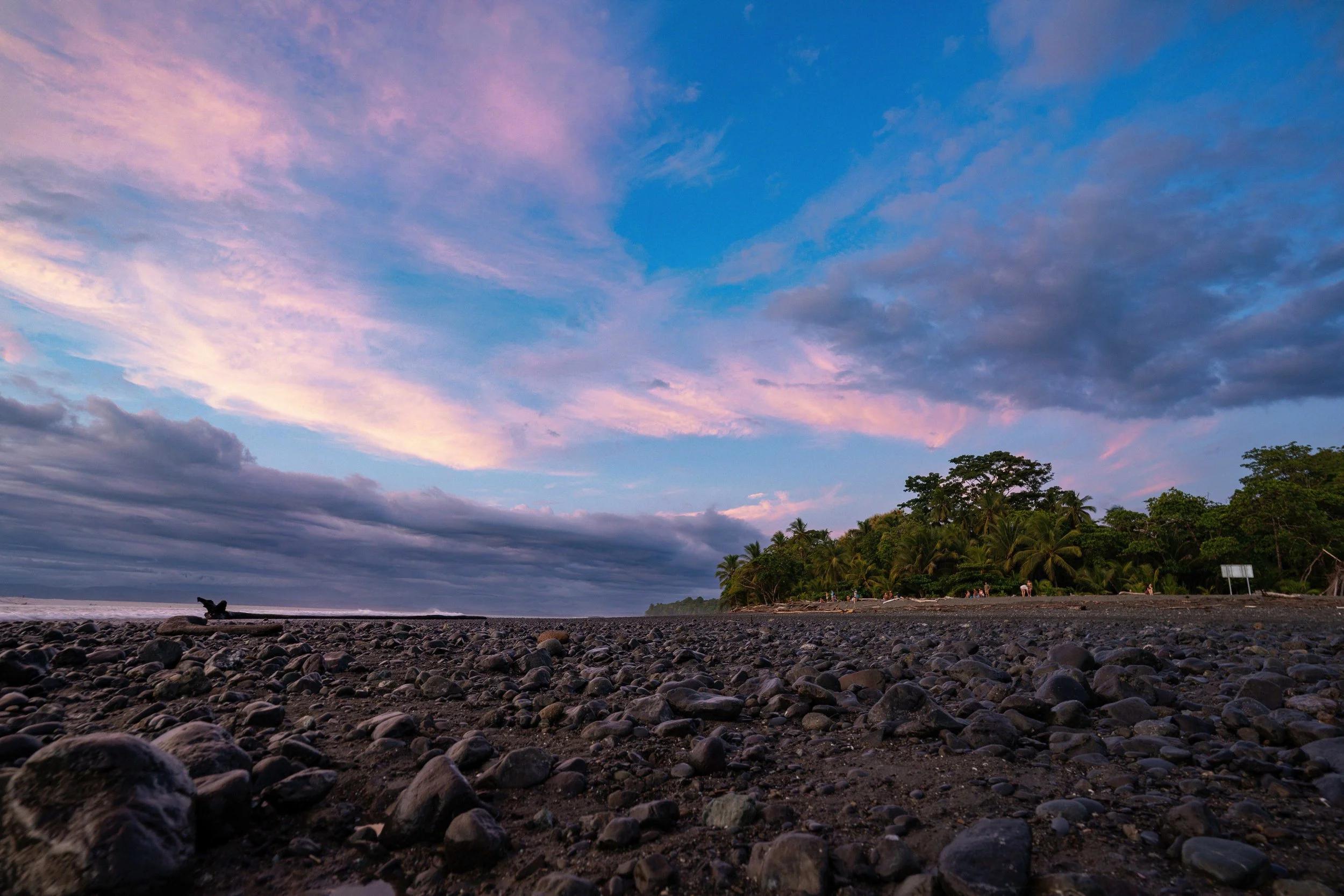A beach with rocky shoreline and a distant tree-lined coast, sky with pink and blue clouds at sunset.
