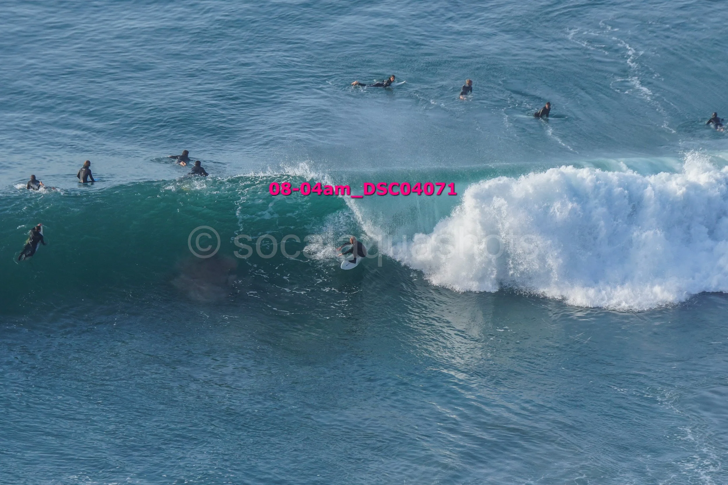 Surfers in wetsuits riding and surfing large ocean waves, with some floating and paddling in the water.