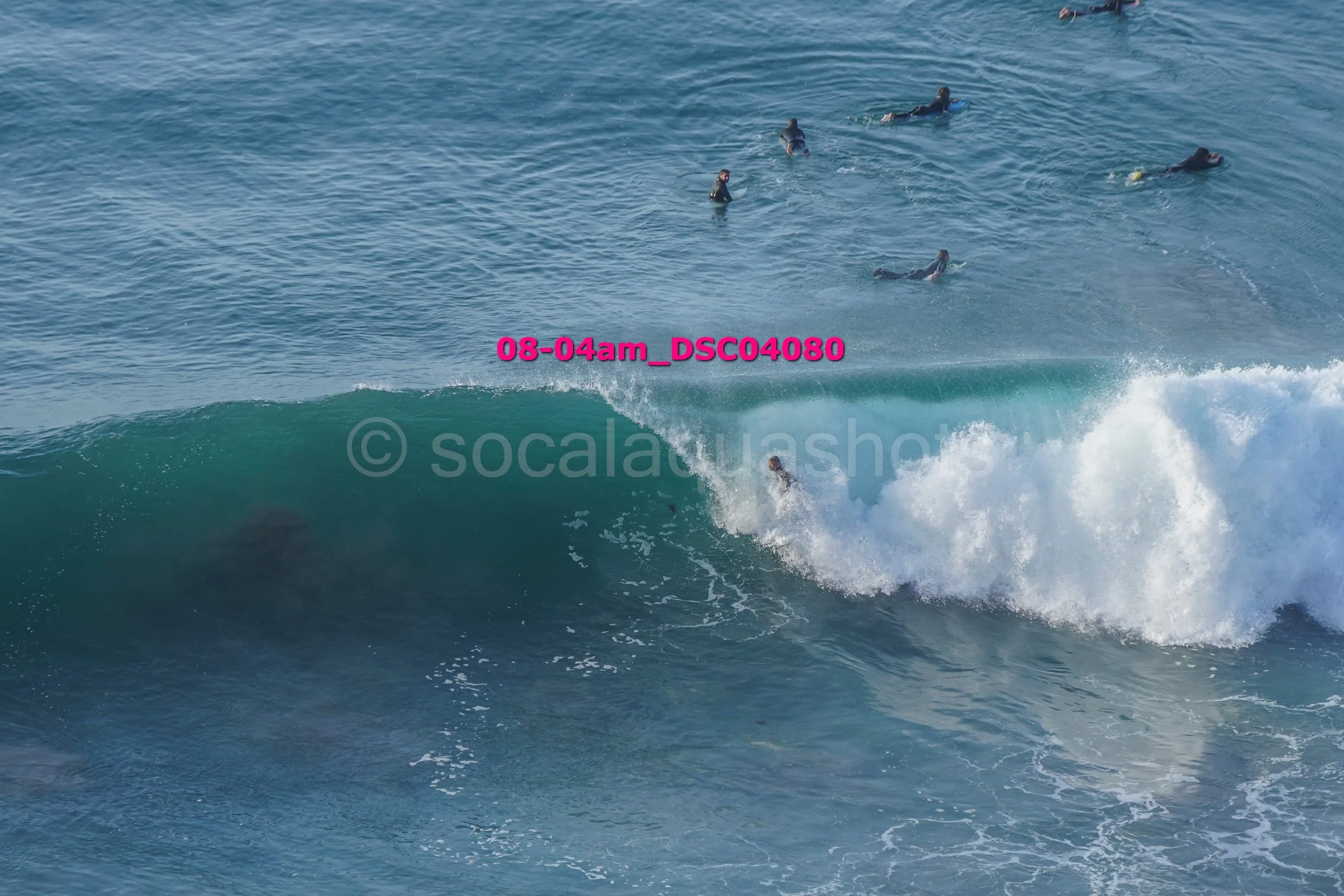A surfer riding a large wave in the ocean, with several other surfers floating in the water nearby.