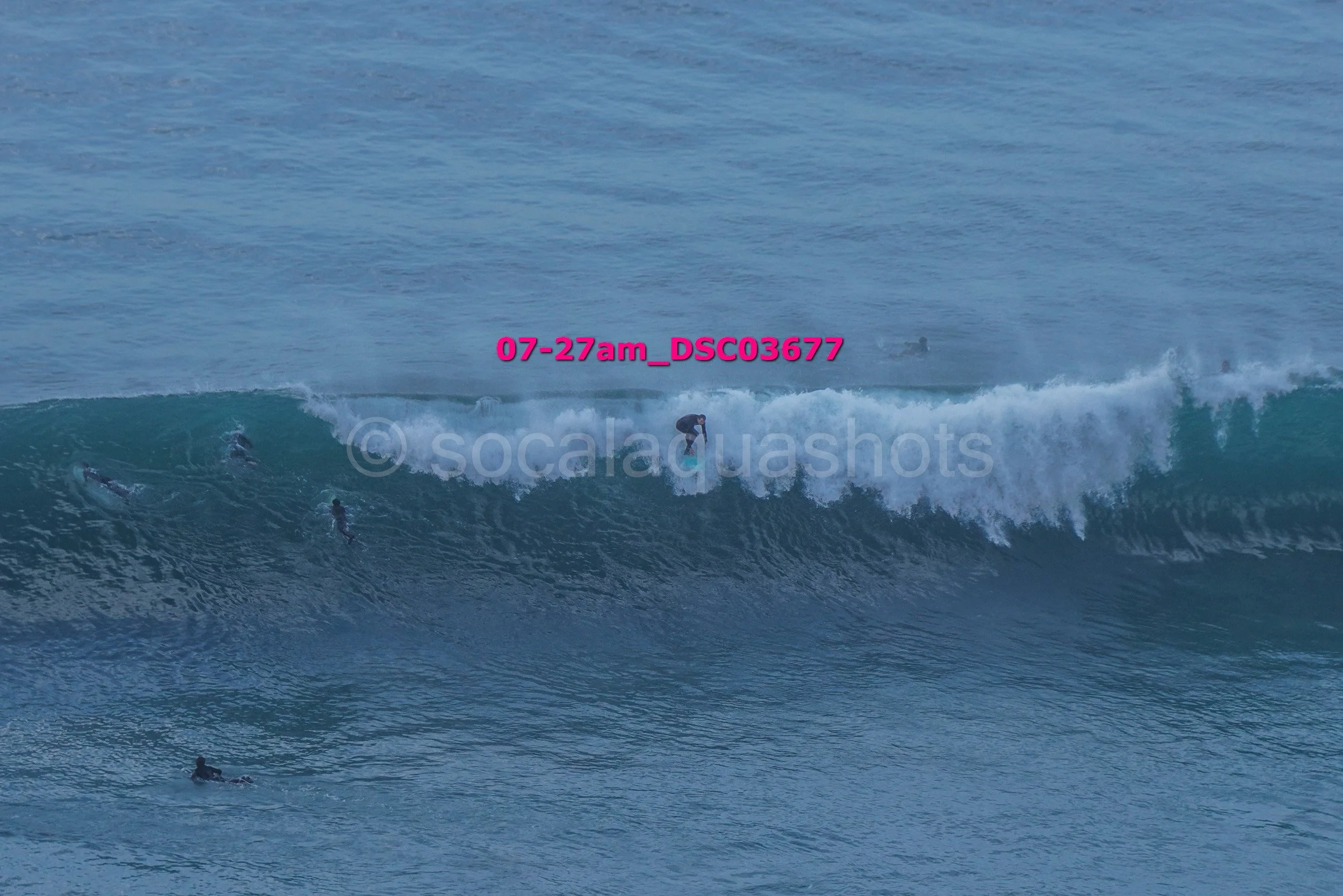 A person surfing on a large ocean wave with several other surfers visible in the water.