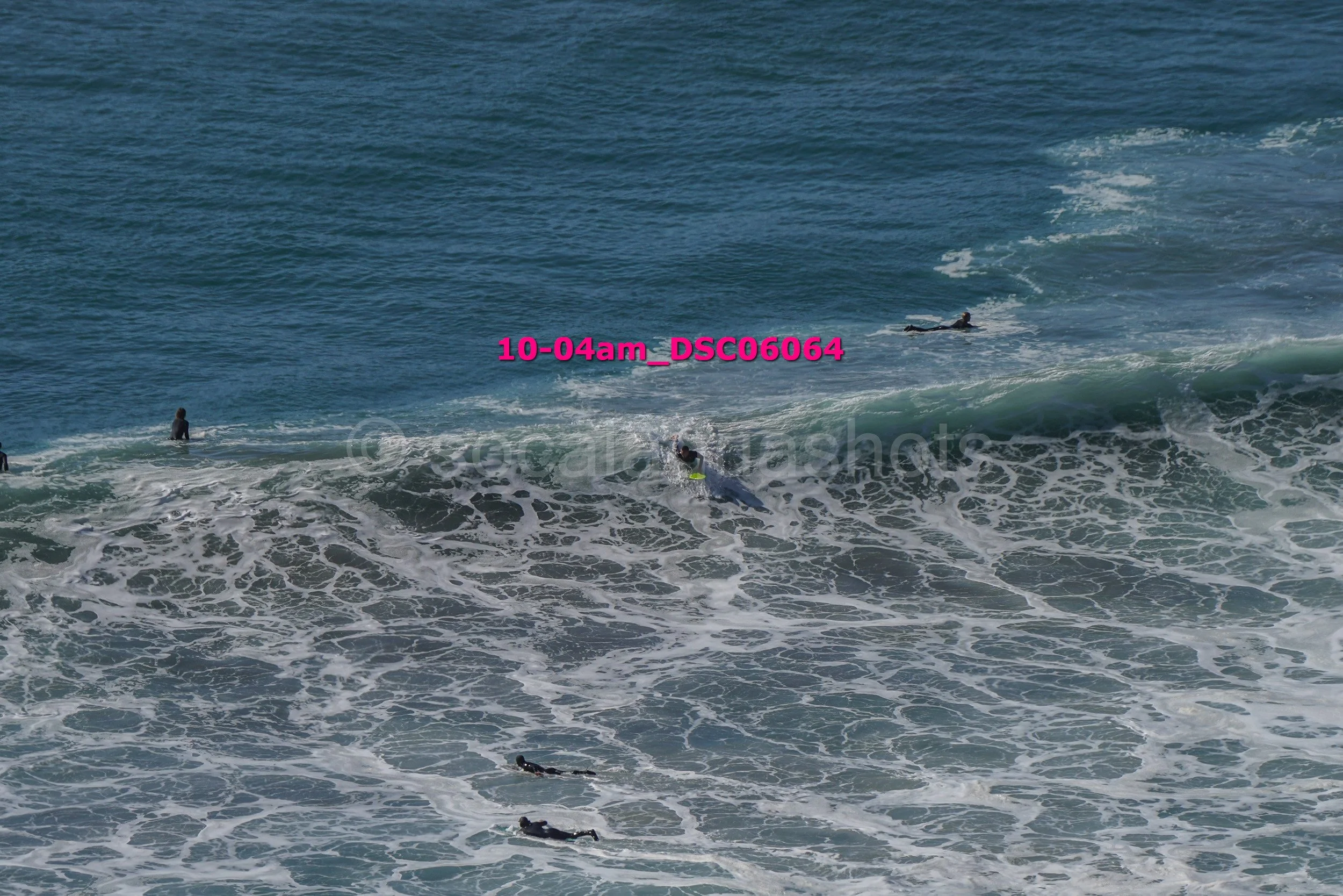 Several surfers in wetsuits riding and waiting for waves in the ocean, with some partially submerged and others lying on surfboards.