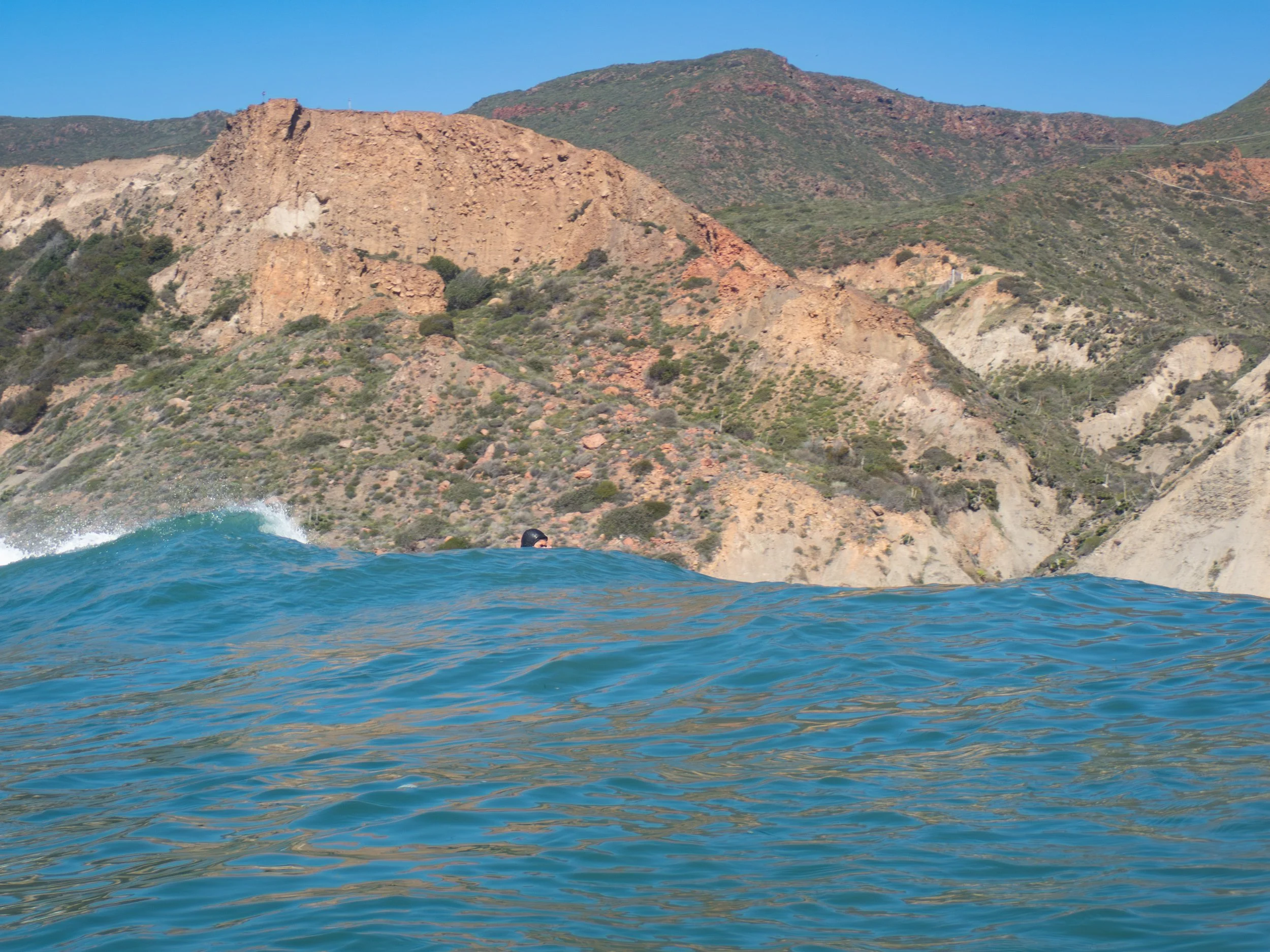 Ocean waves with a distant hillside with sparse vegetation under a clear blue sky.