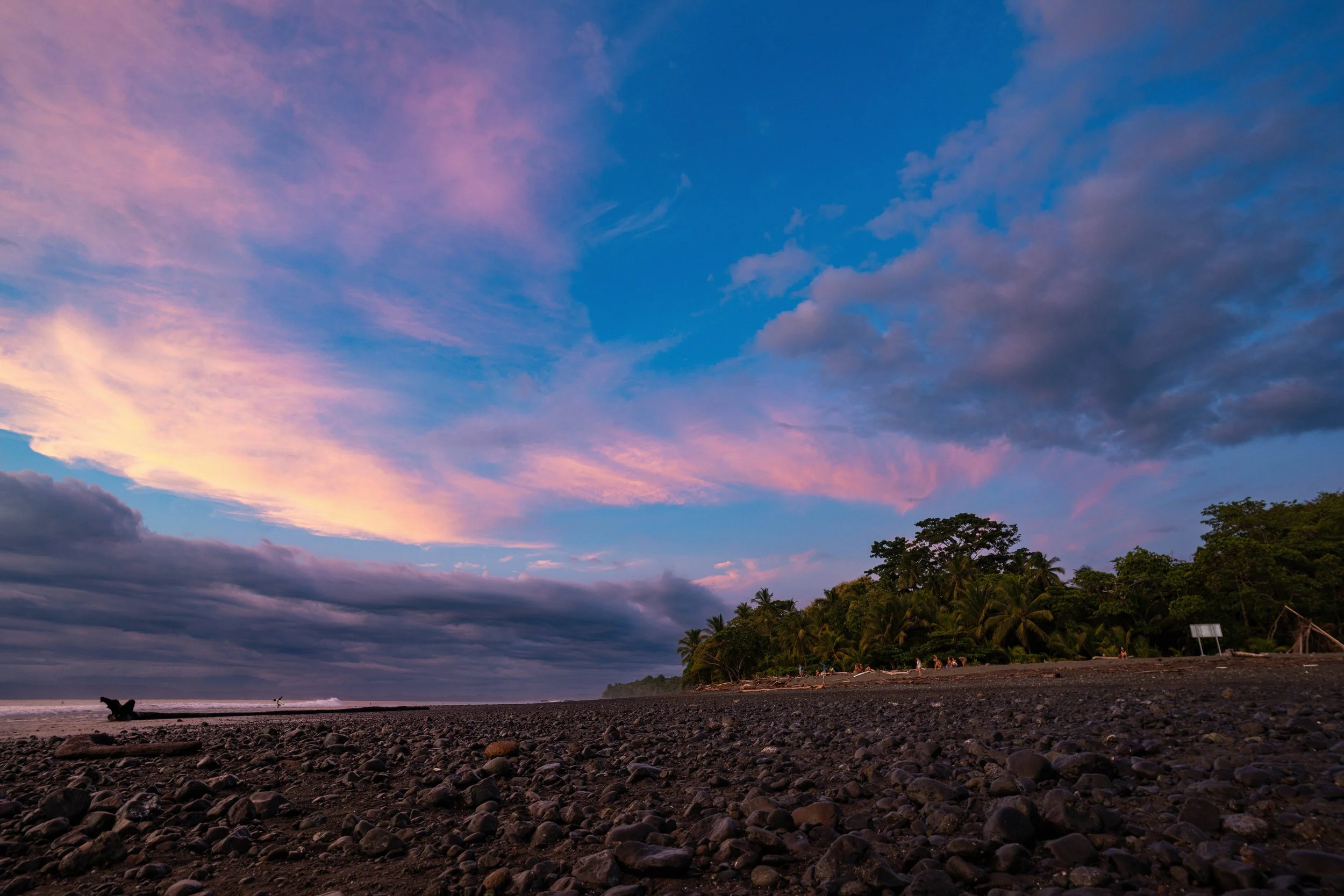 A serene beach scene at dusk with a colorful sky featuring pink and purple clouds, a silhouette of trees, and a rocky shoreline in the foreground.