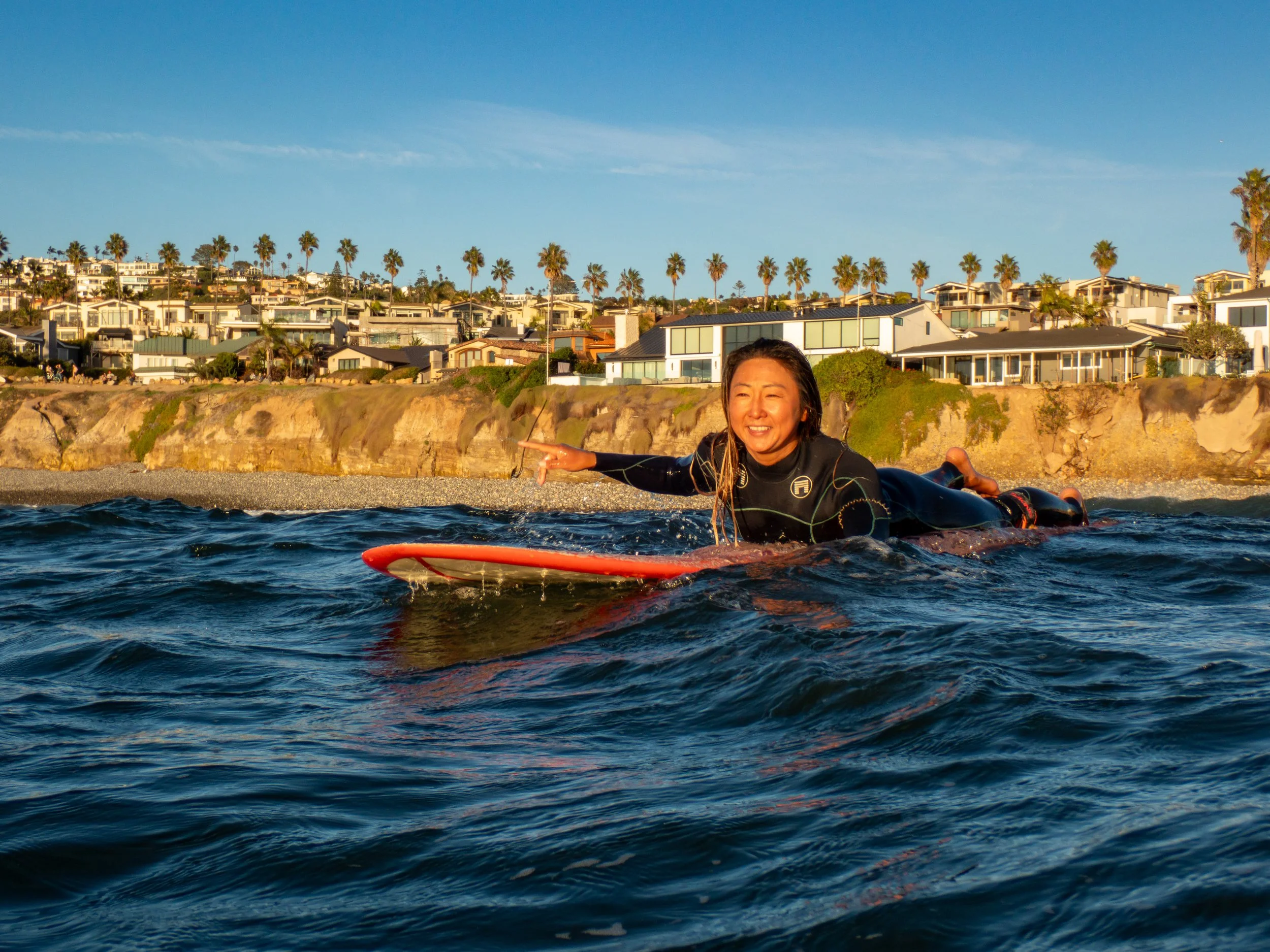 A woman is lying on a surfboard in the water, smiling and pointing, with a coastal residential area and palm trees in the background during sunset.