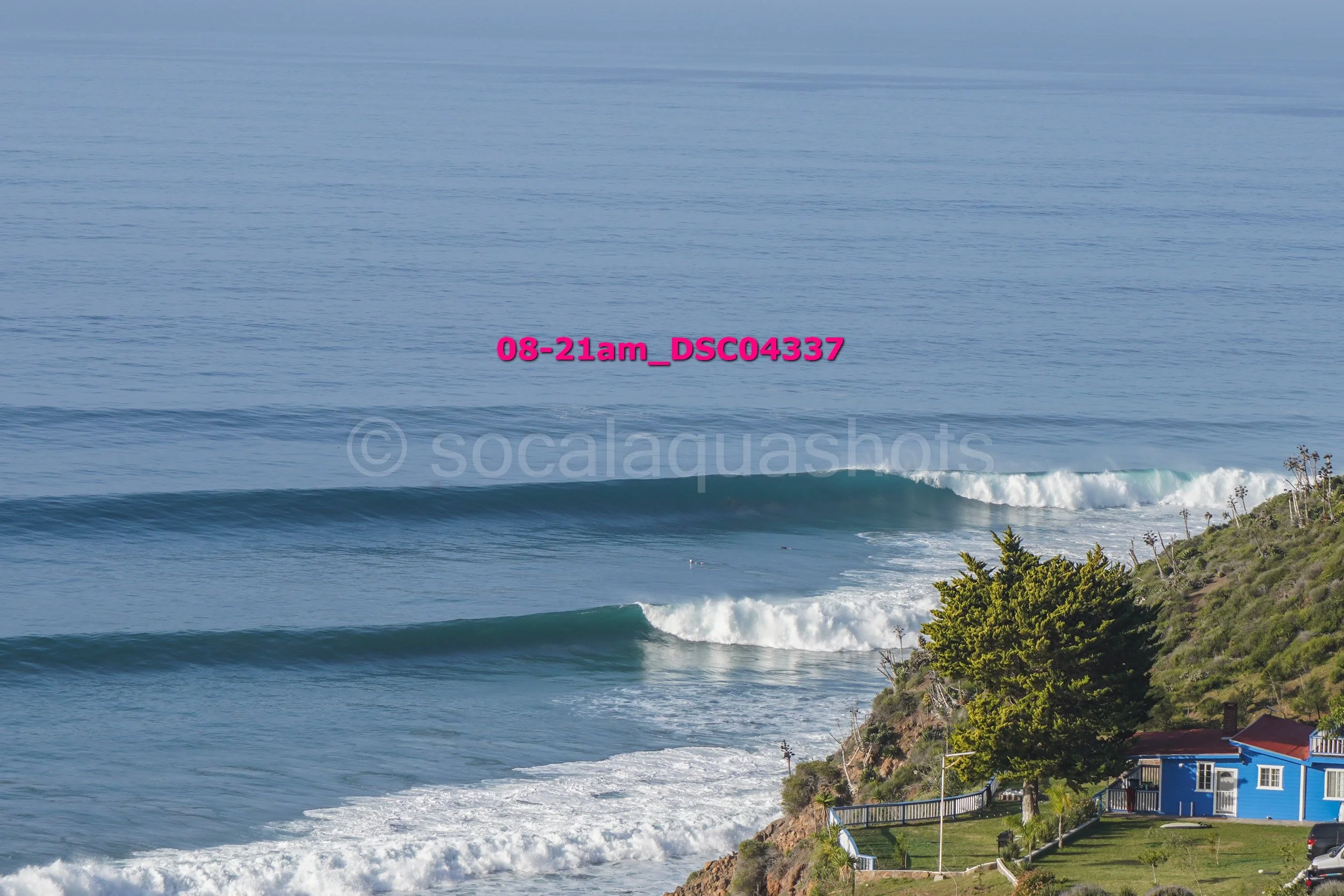 Ocean waves crashing near a coastal hillside with a blue house and trees