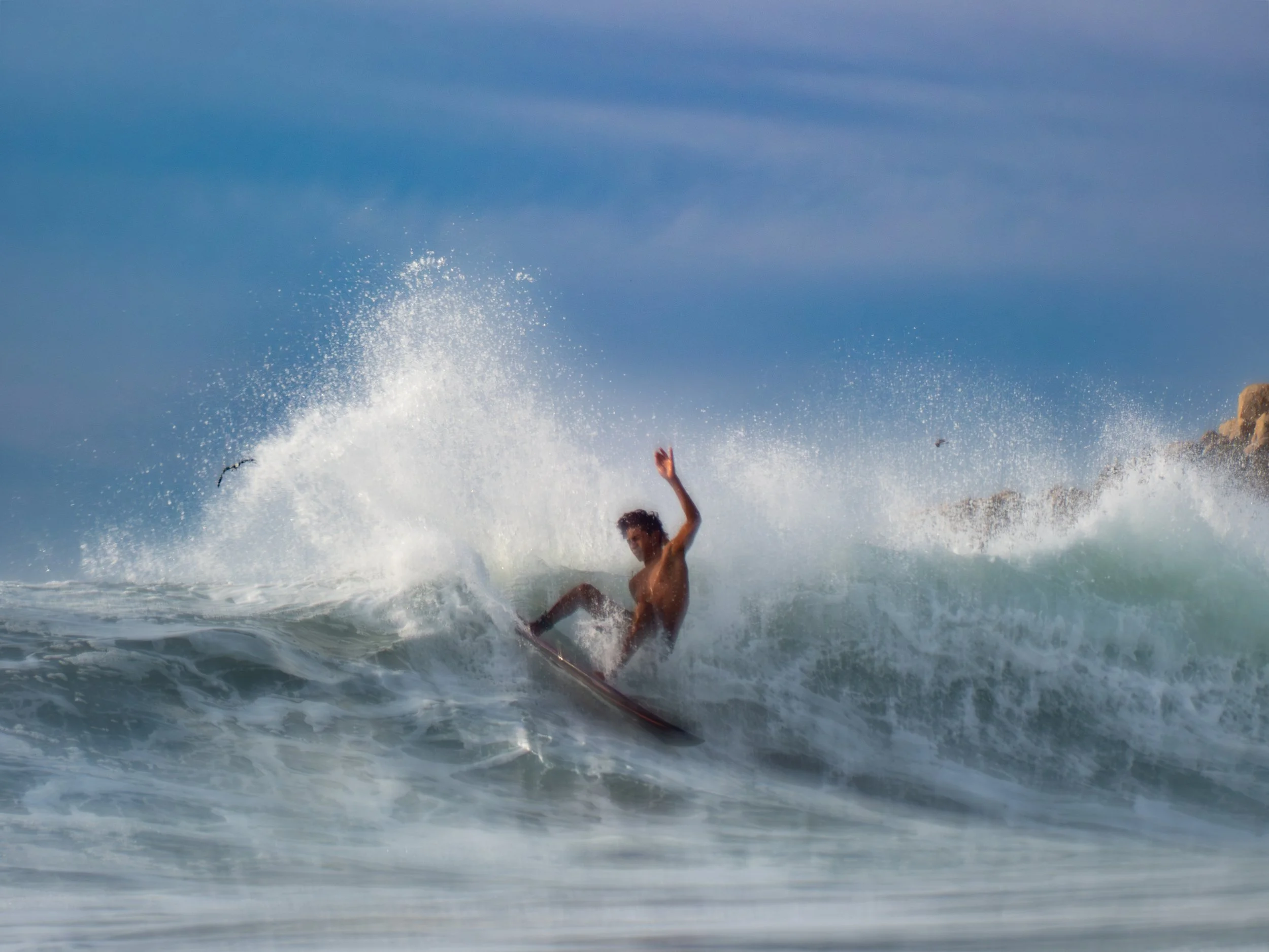 A person falling into the ocean waves after being hit by a large splash, with arms raised and water splattering high in the air under a partly cloudy sky.