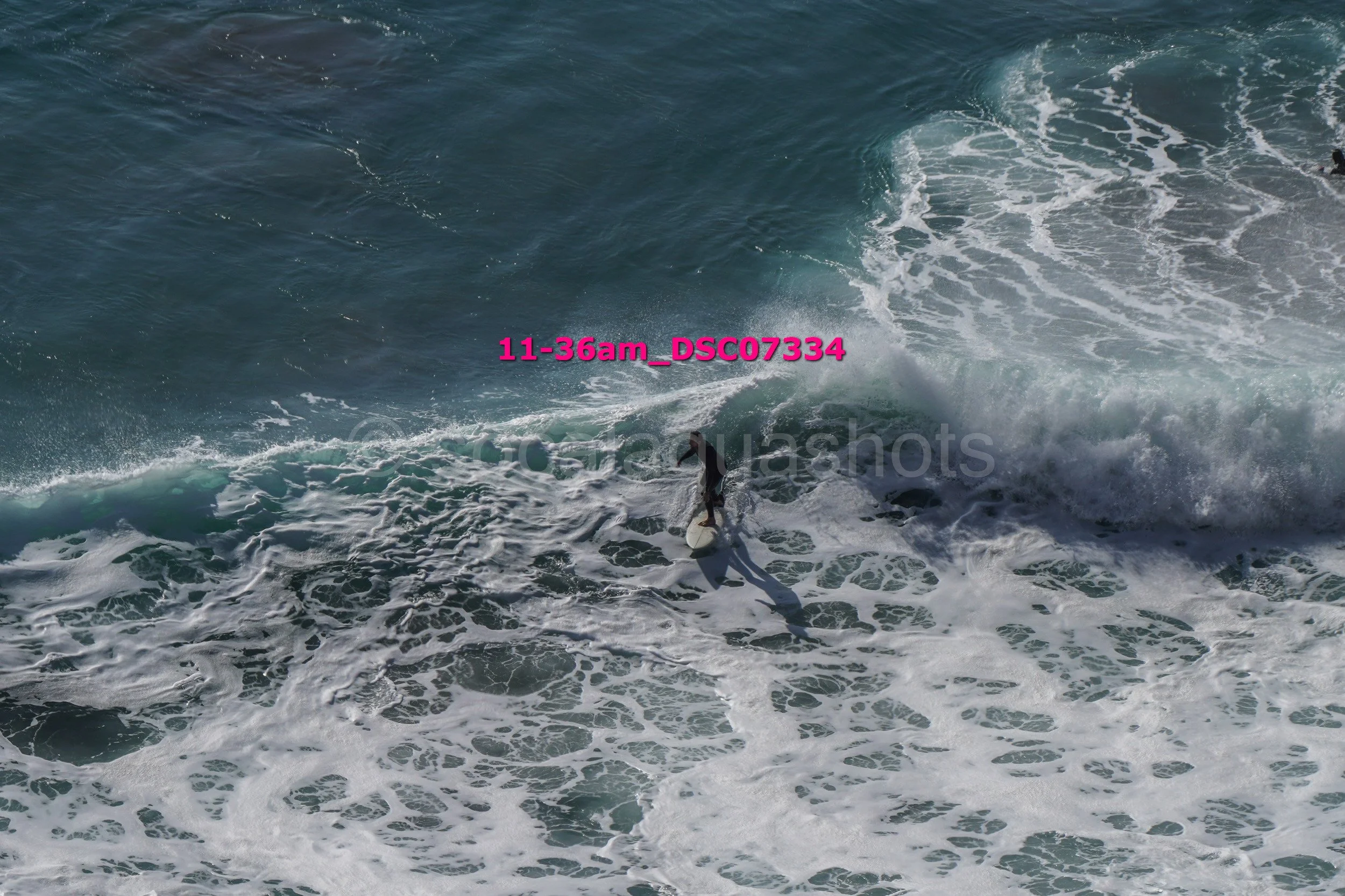 A surfer riding a wave in the ocean with white foamy water, viewed from above.