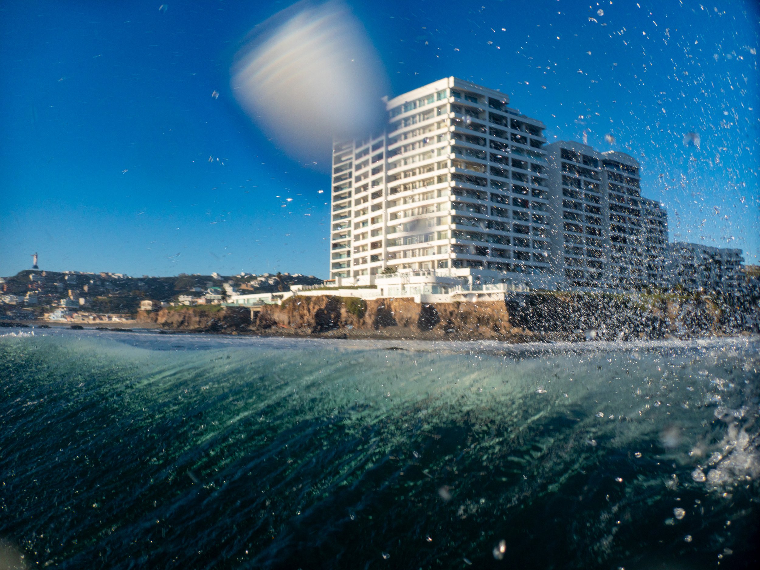 A large, white modern building near the coastline with waves splashing in the foreground, blue sky, and sunlight reflecting on the water.