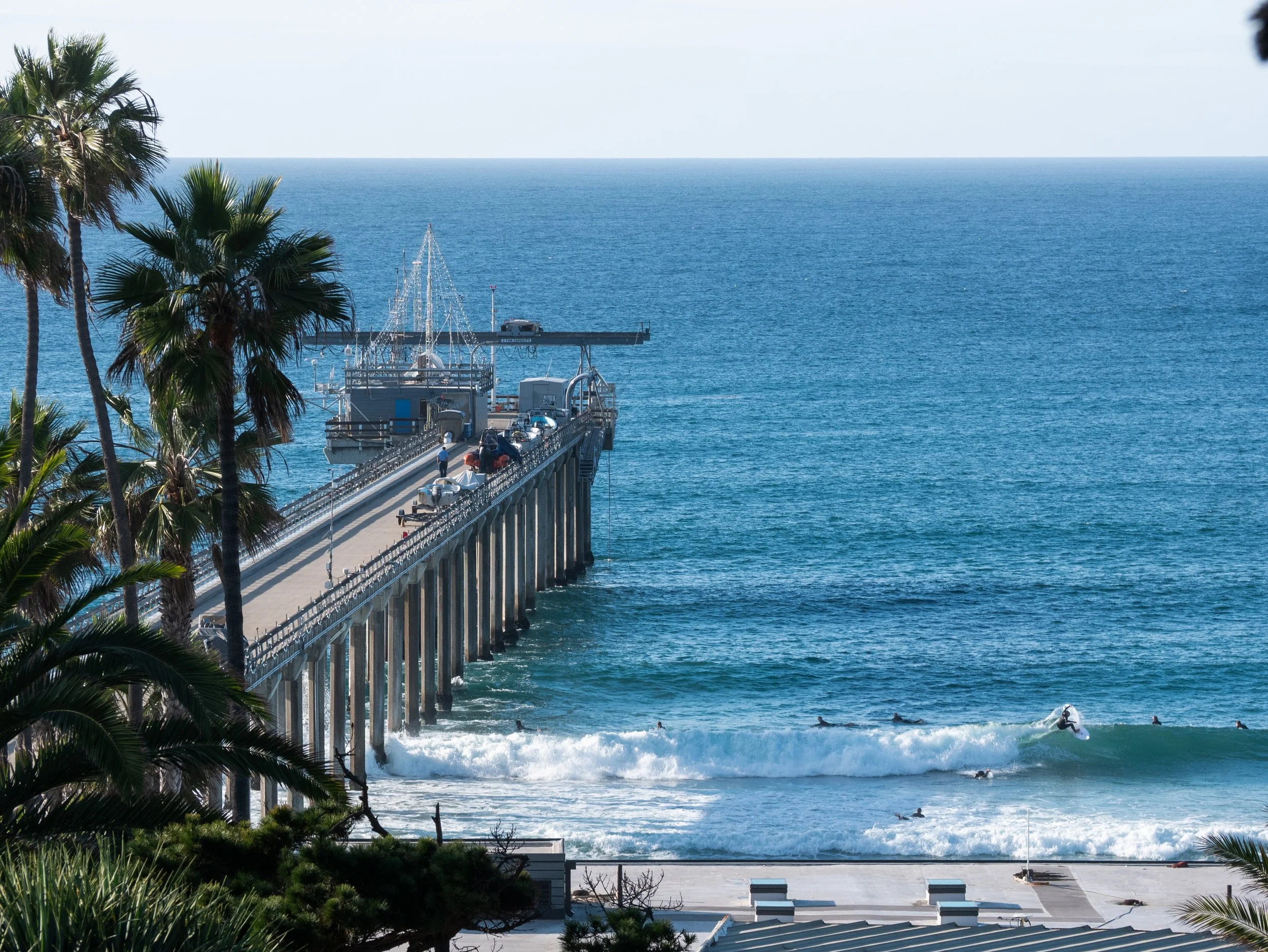 A long pier extending into the ocean with a few people standing on it, surrounded by palm trees, with surfers riding waves in the water and boats docked at the pier.